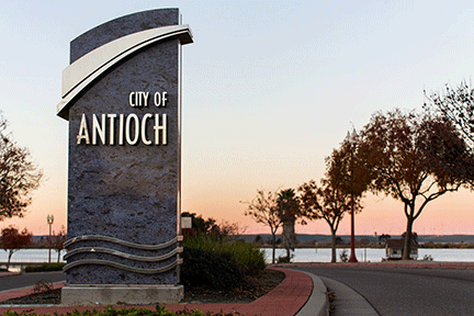 City of Antioch welcome sign near waterfront with trees and a curved sidewalk at sunset.
