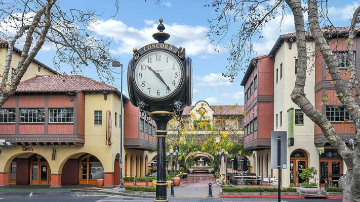 Public square with a decorative street clock showing 5:55, surrounded by trees, buildings, and outdoor seating areas.