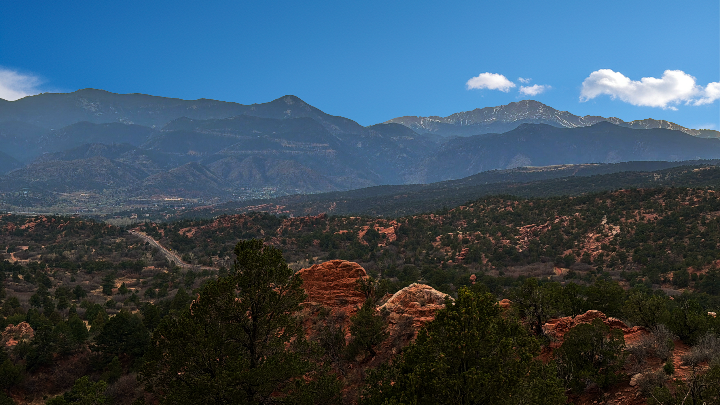 Mountain landscape with red rocks and green trees in the foreground, rolling hills, and a range of mountains in the background under a blue sky with a few clouds.