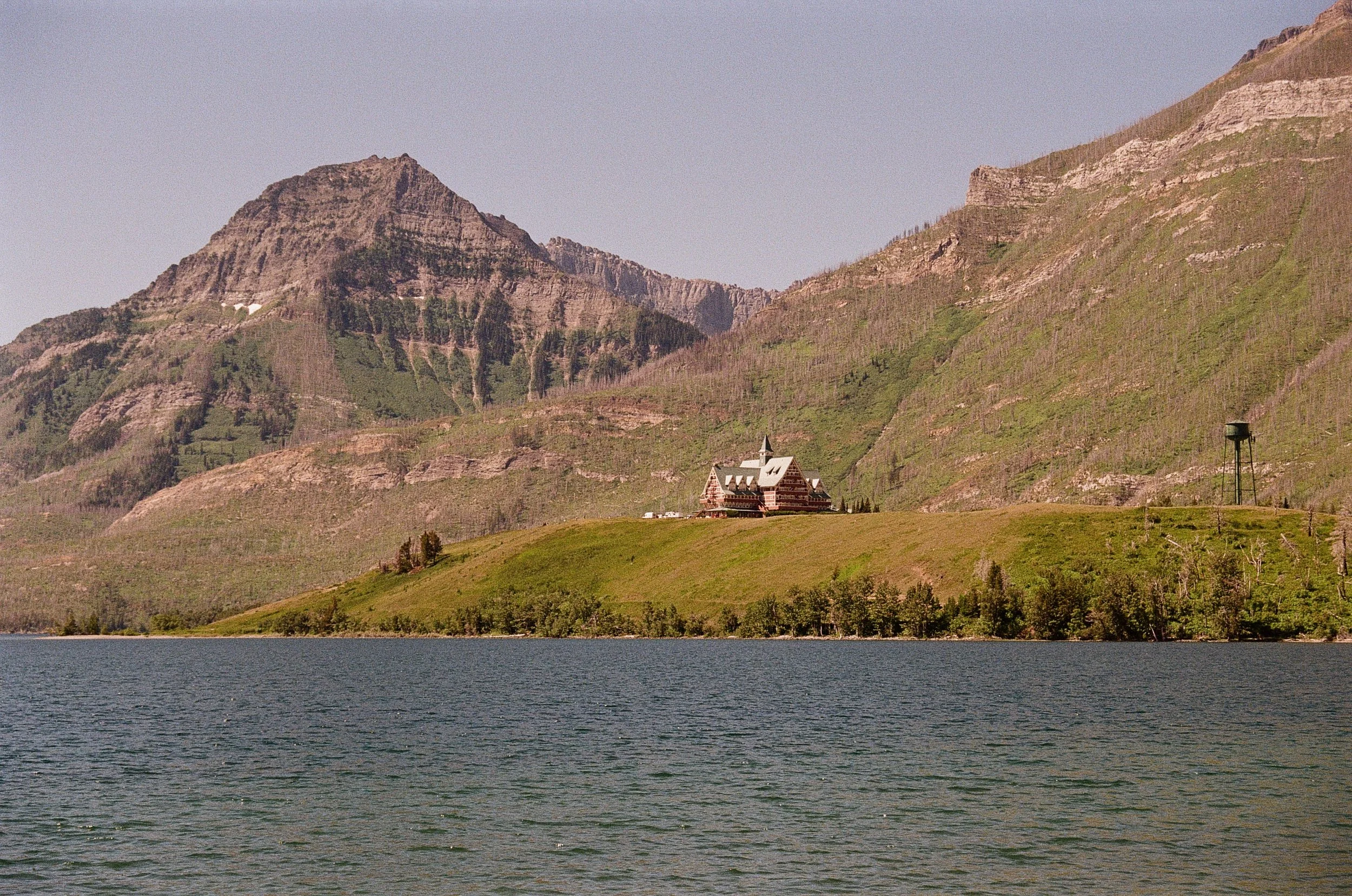 A large body of water in the foreground, with a grassy hill and trees leading up to a church or castle-like building on the hill. In the background, tall mountains with some greenery and rocky peaks are visible under a clear sky.