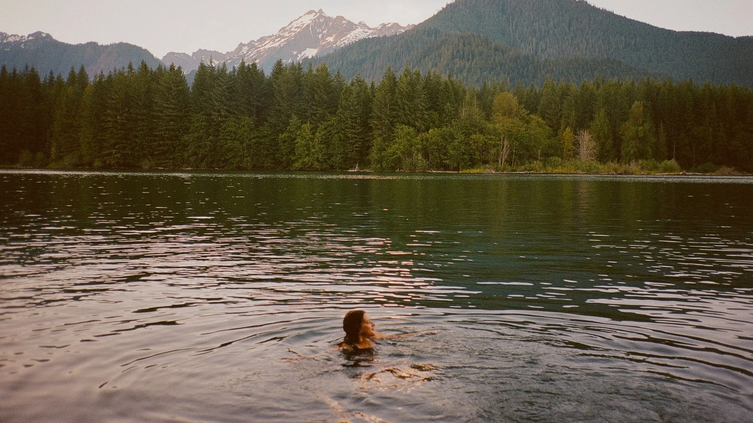 A person swimming in a lake surrounded by green trees and mountains with snow-capped peaks in the background.