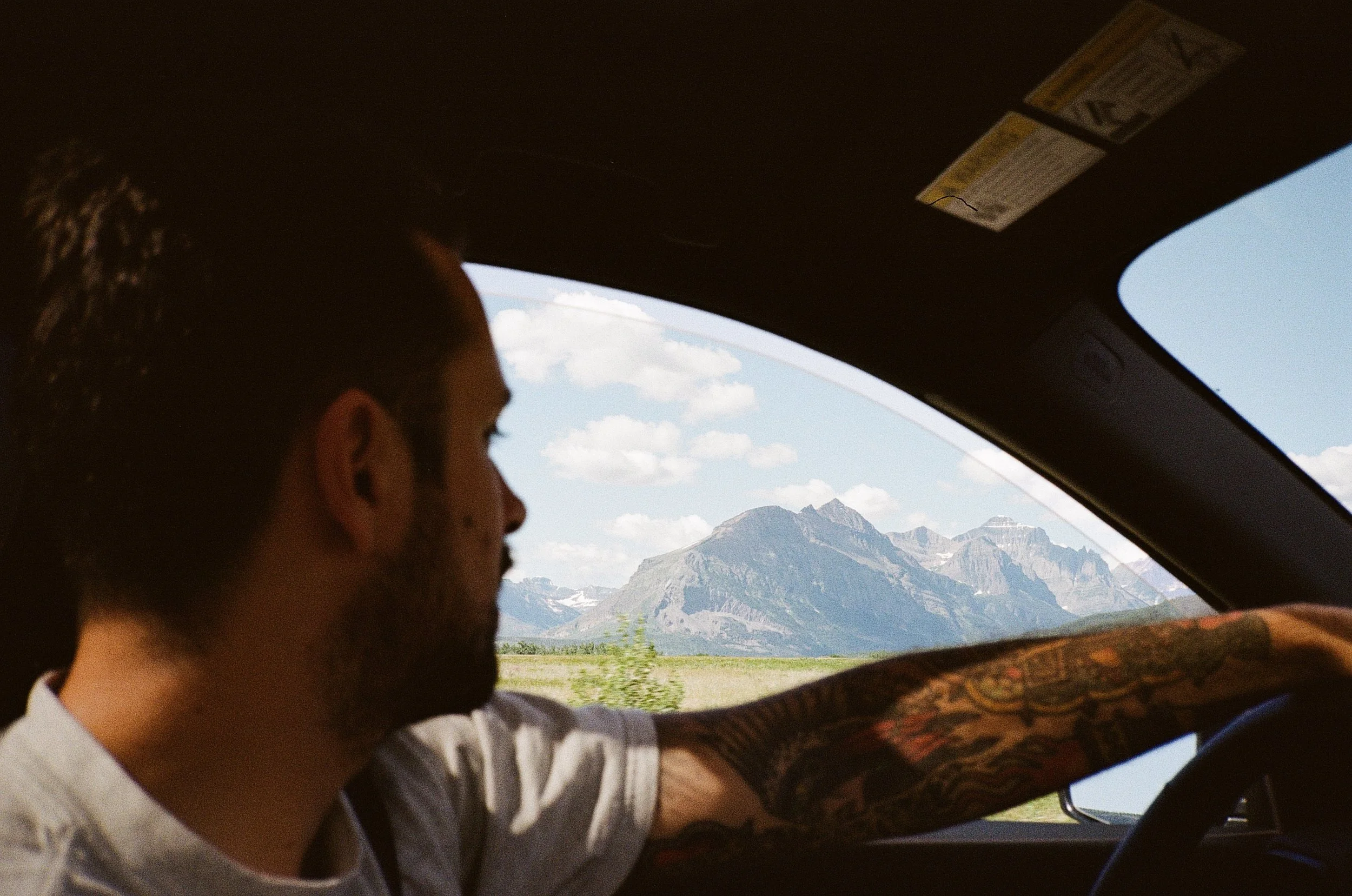 A man with tattoos driving a vehicle with a mountain landscape outside the window.