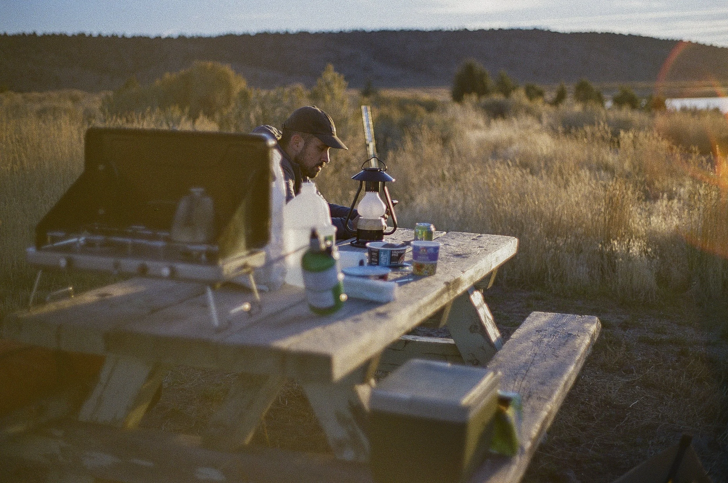 A man sitting at a rustic wooden picnic table outdoors in a field, with a lantern and camping supplies, during sunset with hills in the background.