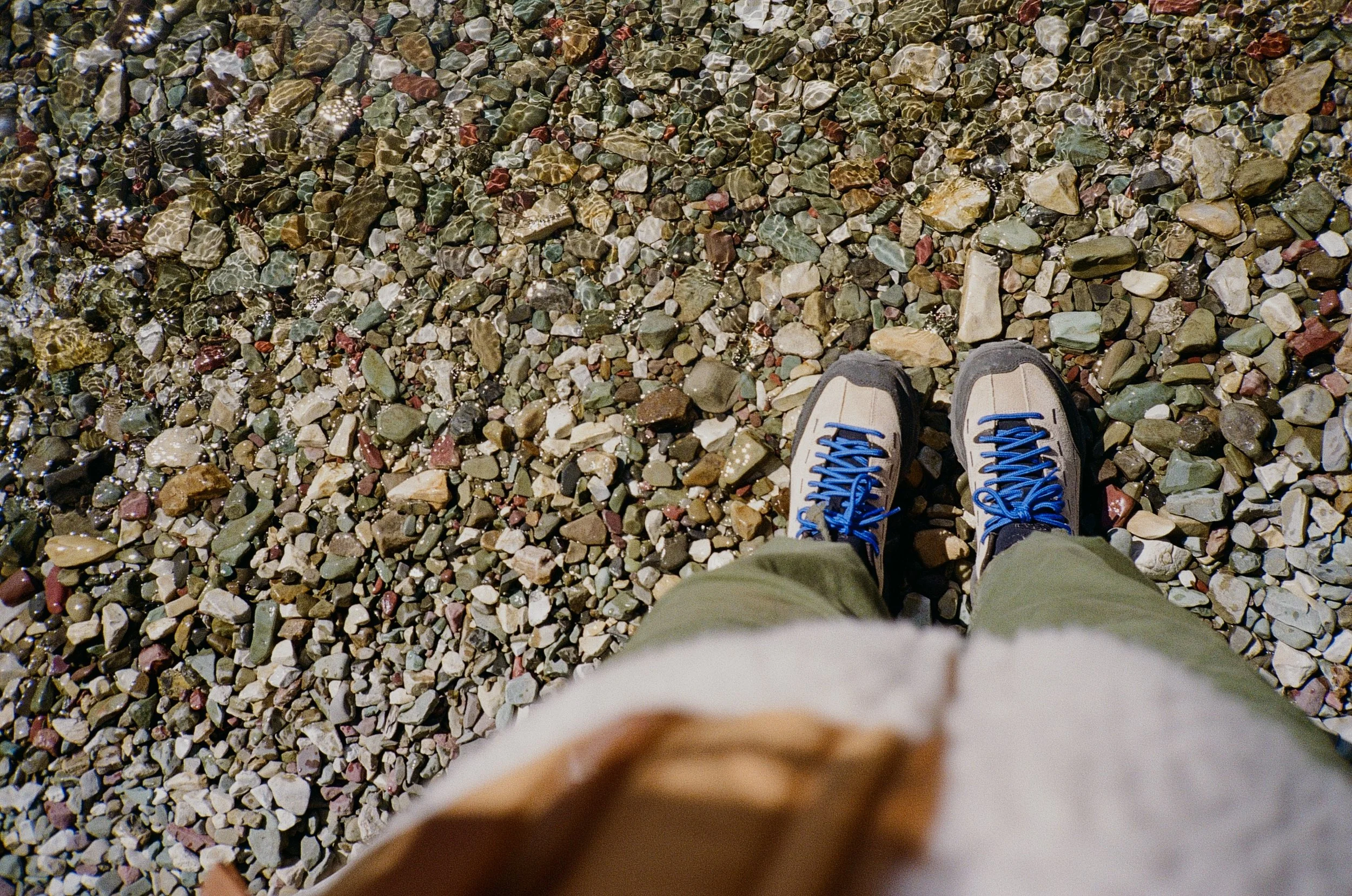 A person's feet in hiking shoes with blue laces standing on a colorful rocky beach with small rocks and pebbles.