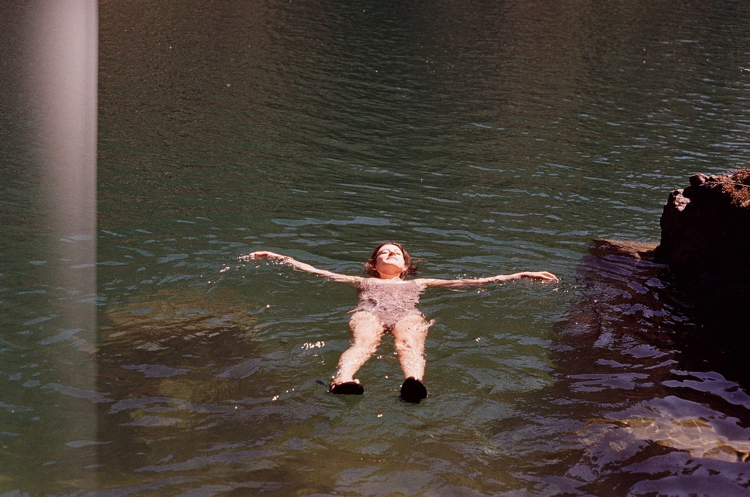 A woman floating on her back in a body of water, with arms outstretched and eyes closed, near a rocky area.