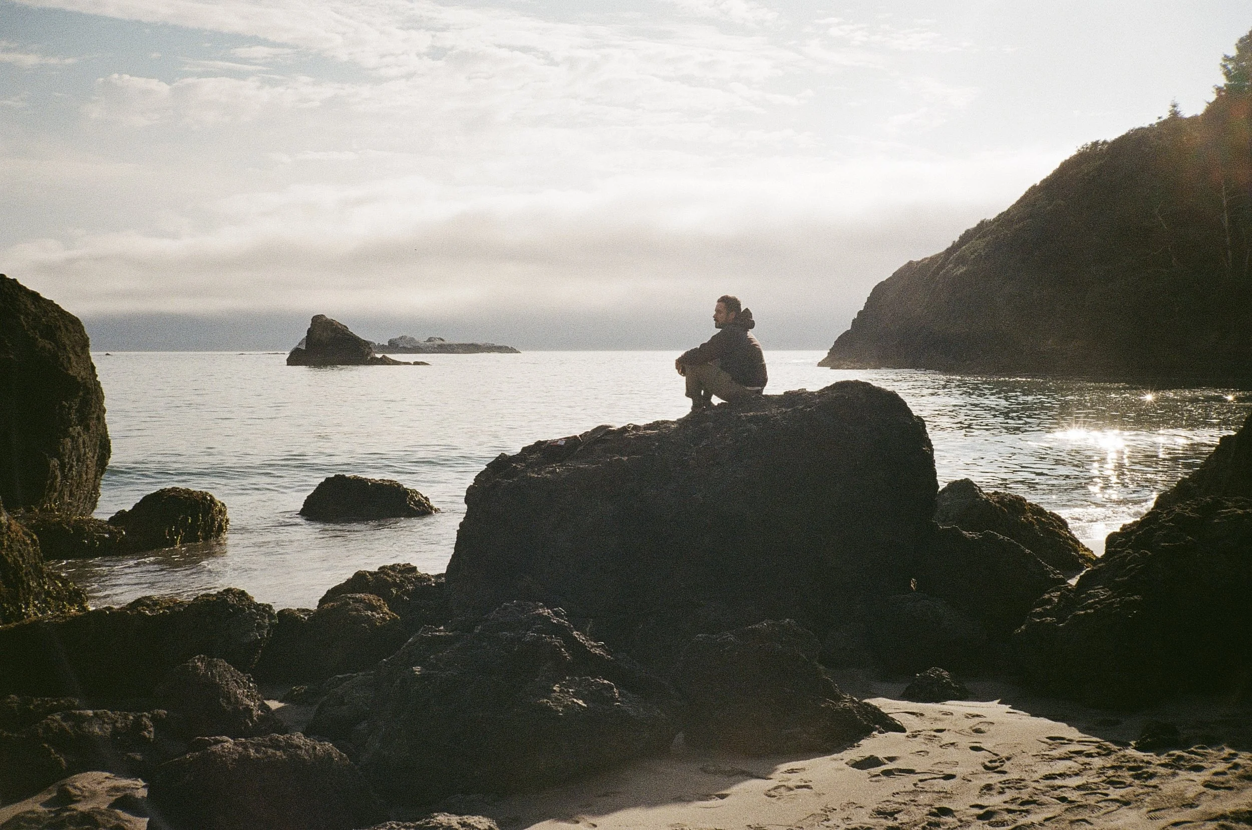 A person sitting alone on a large rock by the shoreline, gazing at the water and distant rocks in a peaceful, natural coastal setting during daytime.