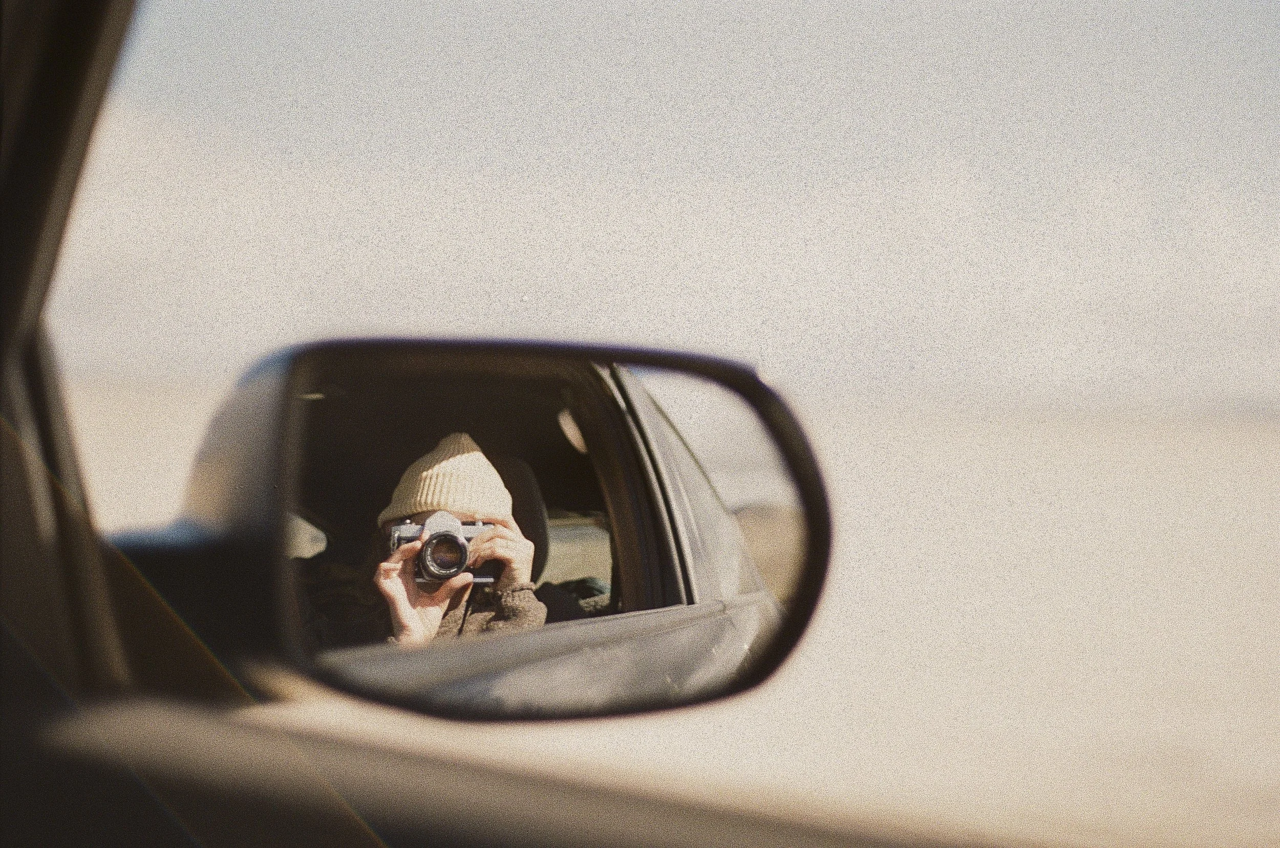 Person with a beige knit hat taking a photo with a camera, visible in the car's side mirror, against a plain sky background.