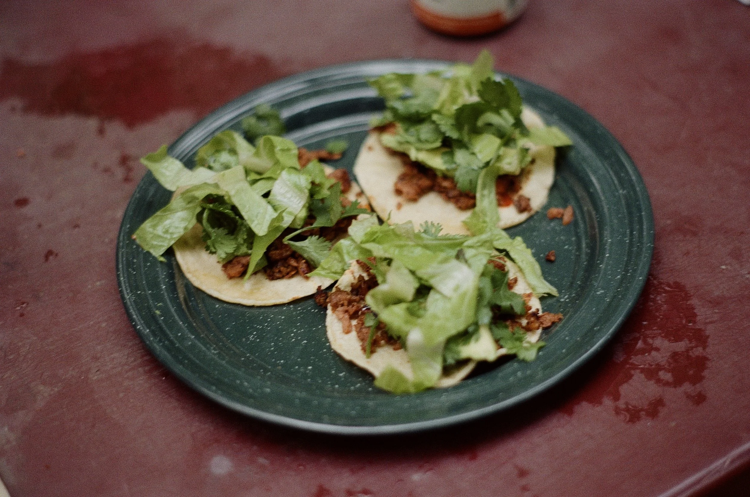 Three tacos on a dark green plate, filled with shredded meat, lettuce, and cilantro, resting on a red table.