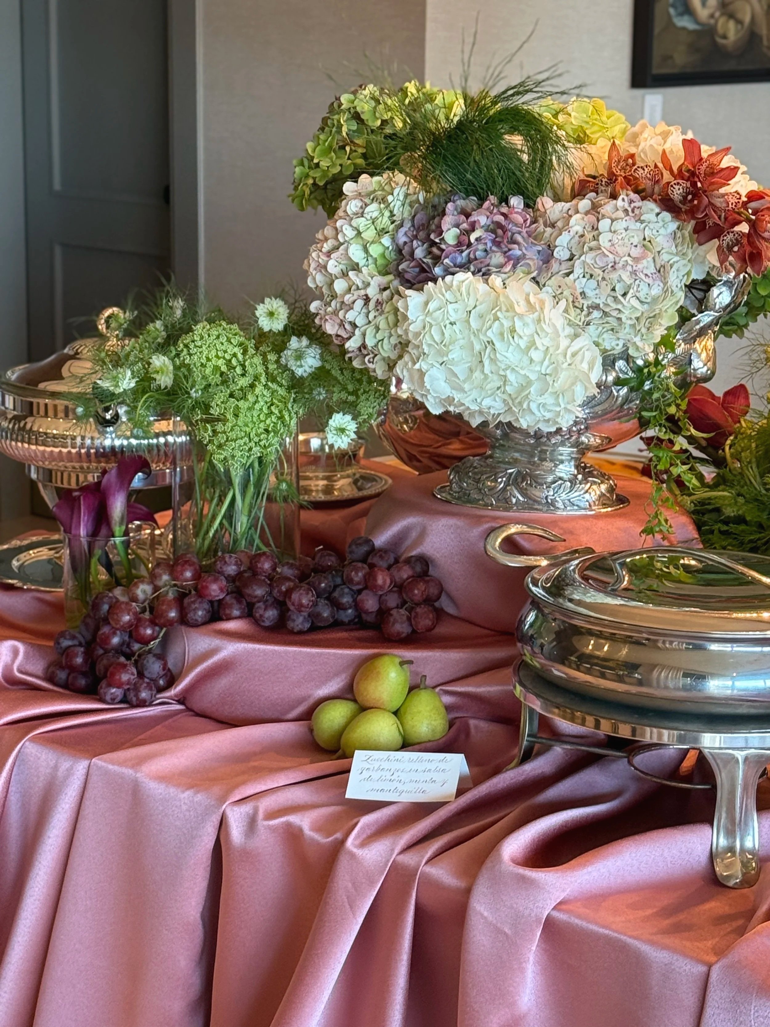 Mesa decorada con un mantel de seda rosa, floreros con flores y uvas, y platos de plata. En el fondo se observa un cuadro y una pared beige.