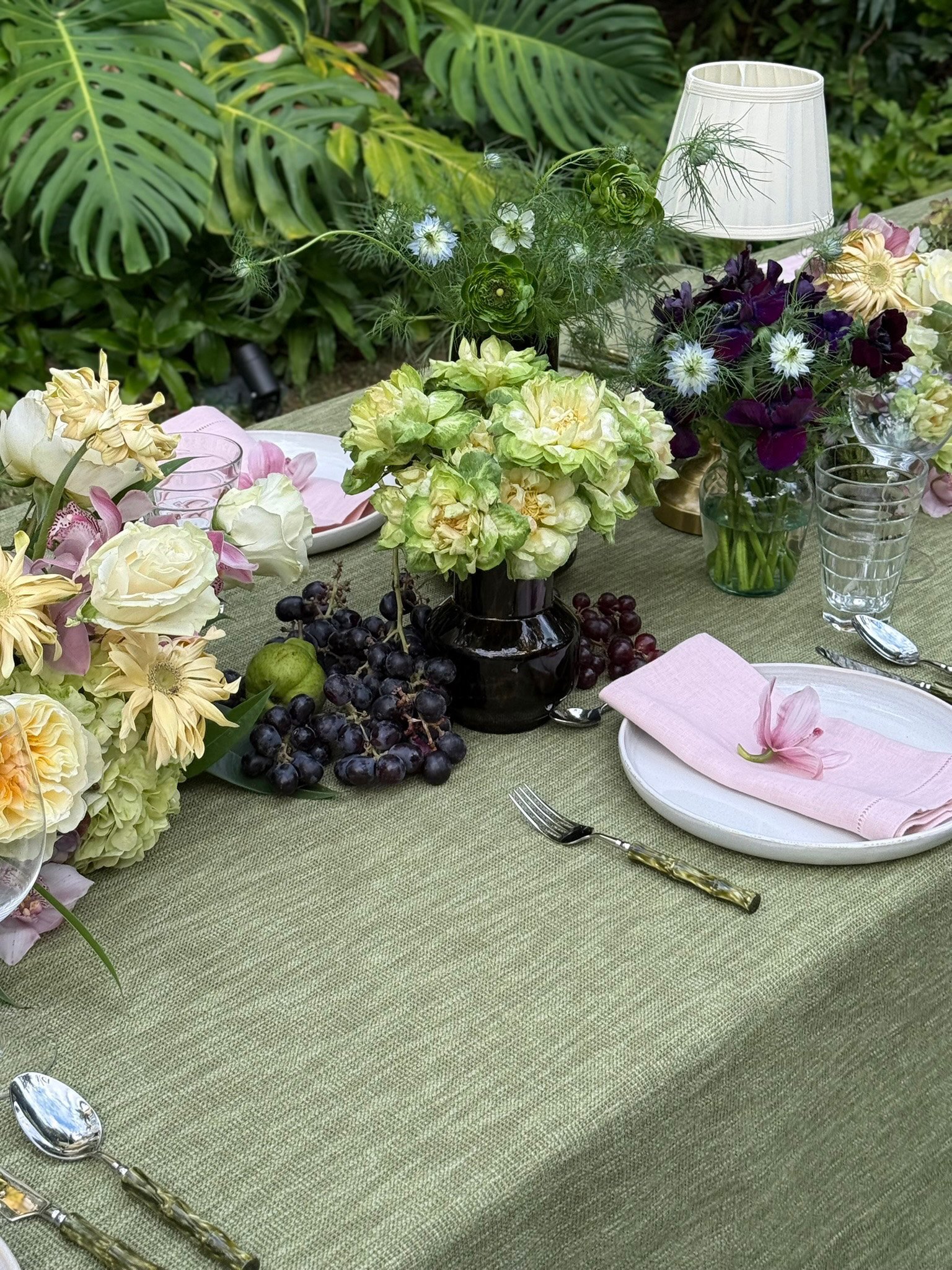Mesa decorada con arreglos florales en jarrones, servilletas rosadas, platos blancos, tenedores y cucharas, con uvas negras y verdes, y una lámpara de mesa en un fondo de plantas verdes.