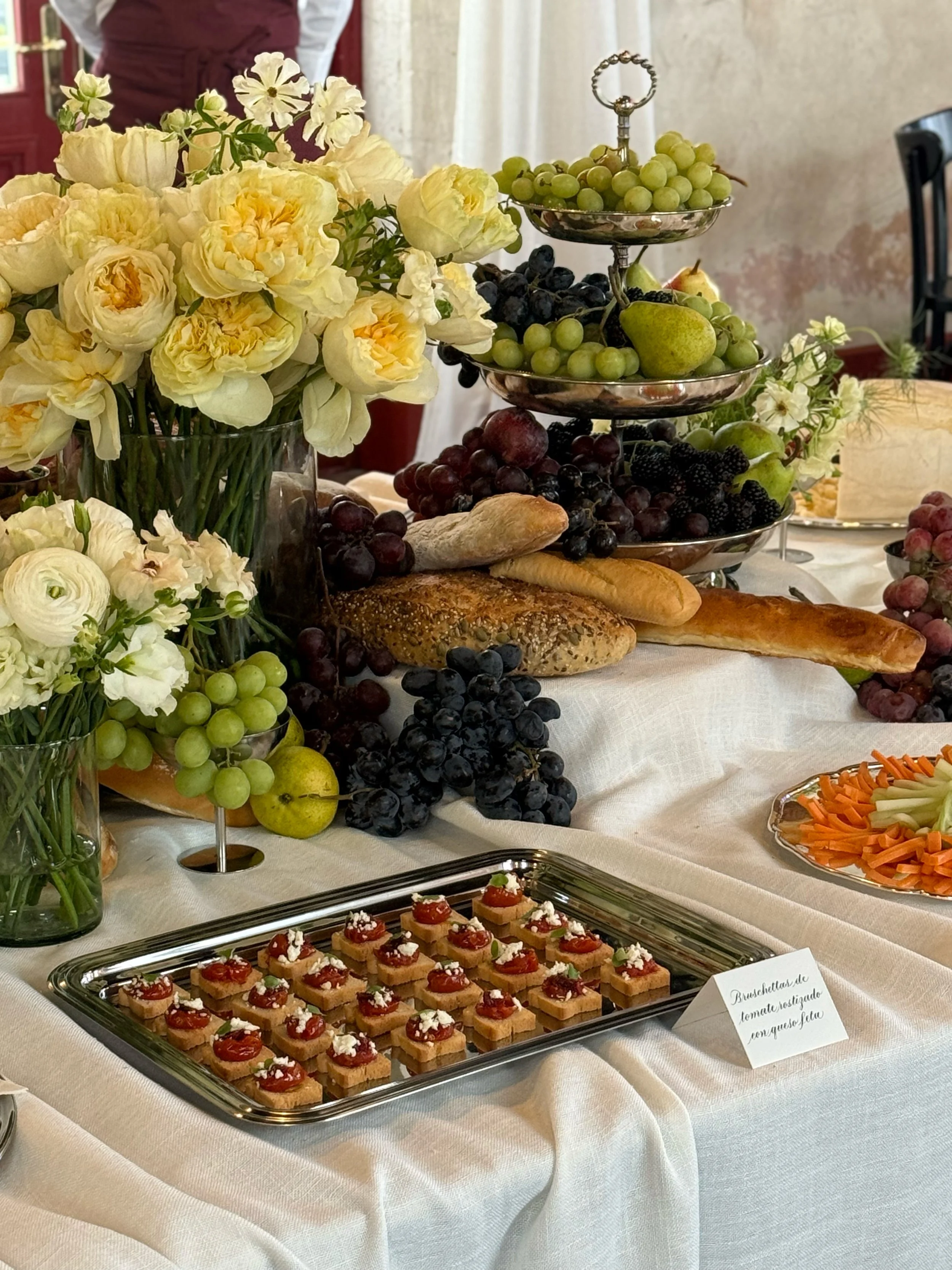 Mesa con variedad de frutas, flores blancas y platos con aperitivos y postres, en ambiente elegante. Incluye uvas verdes y negras, peras, pan, y una bandeja con pequeños bocados decorados con tomate y queso.