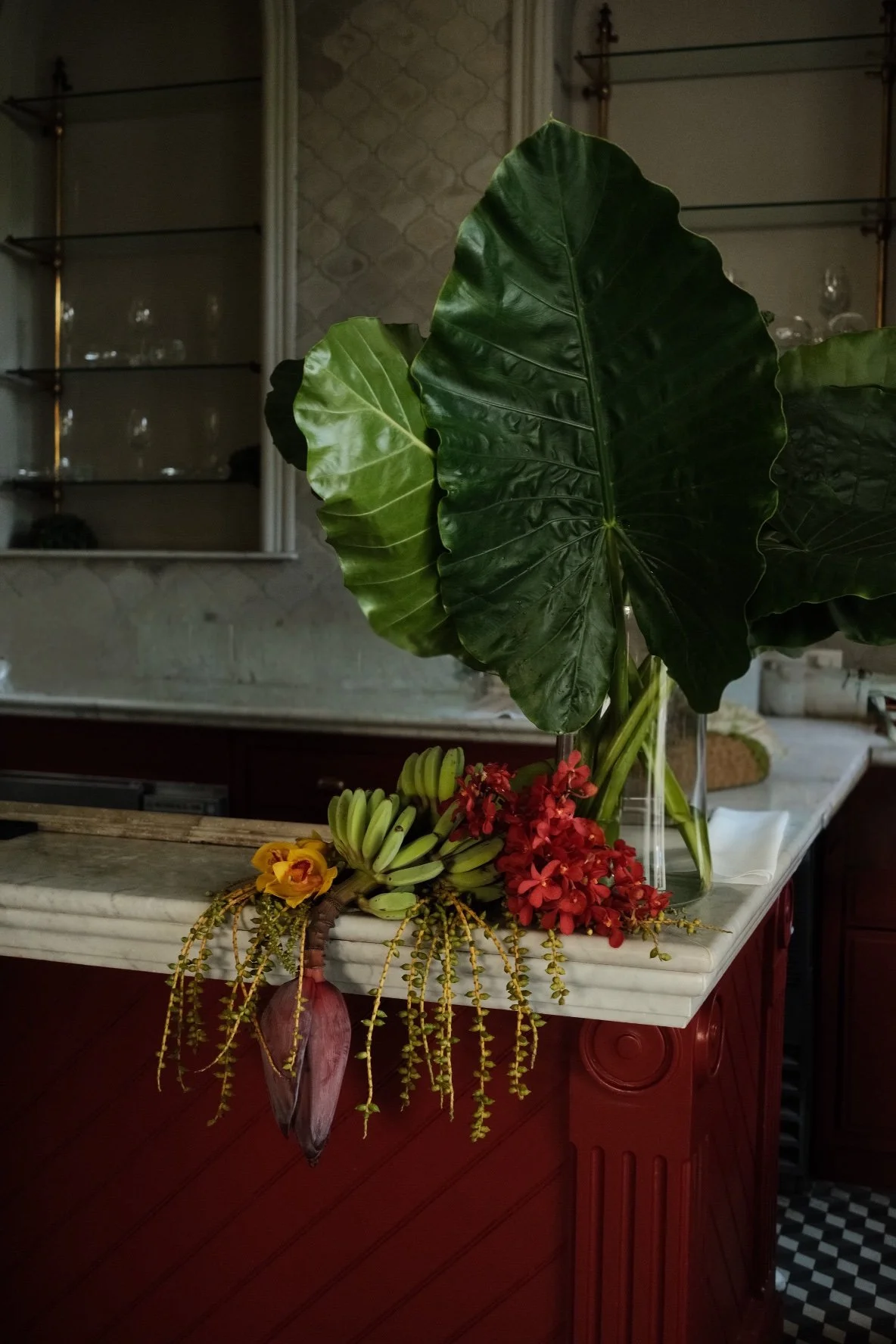 Arreglo floral en una mesa con hojas verdes grandes, plátanos pequeños, flores rojas y amarillas y un florero de vidrio en una cocina o comedor.