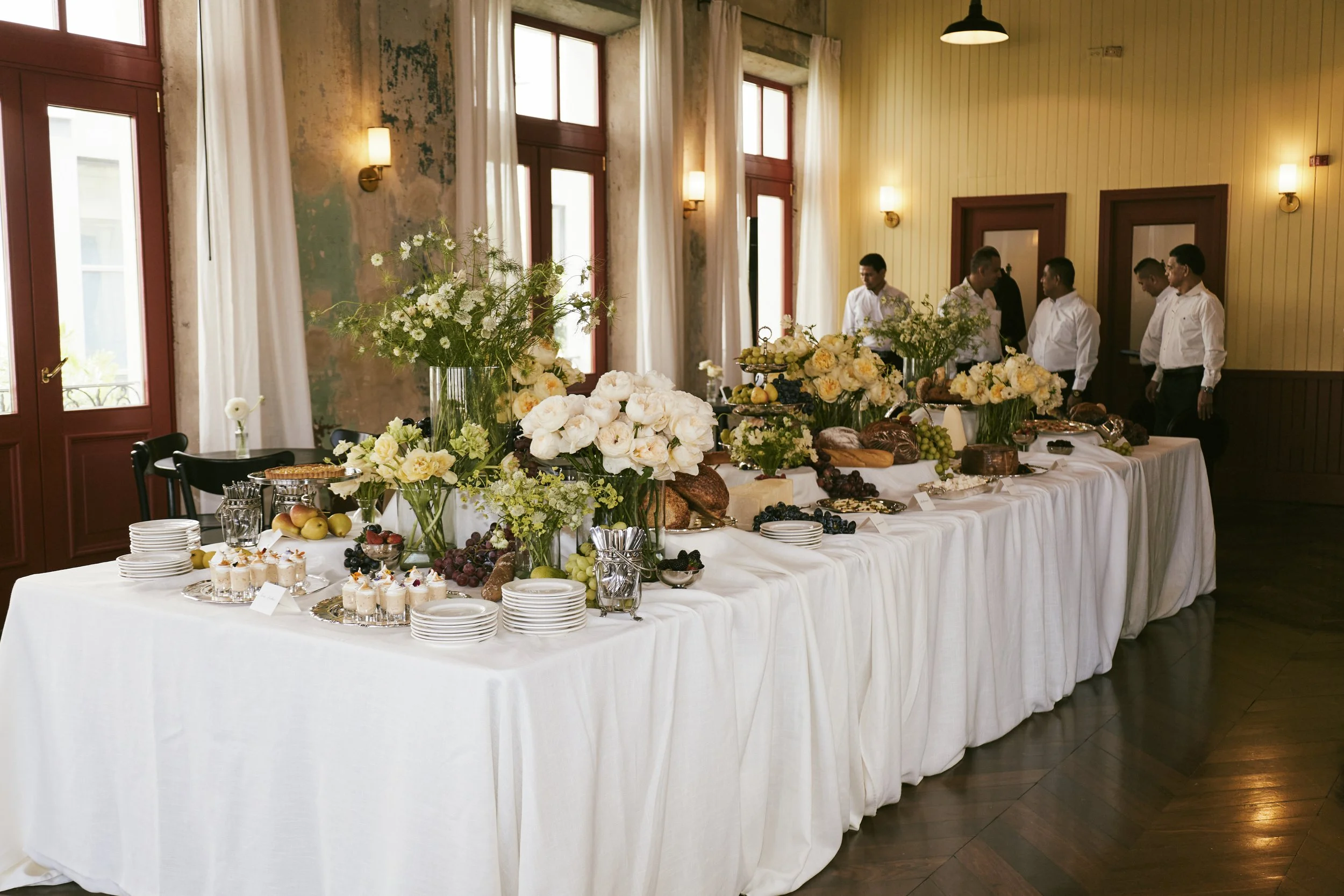 Mesa larga con decoración floral y variedad de alimentos, en un lugar con paredes de madera y personas de traje blanco en el fondo.