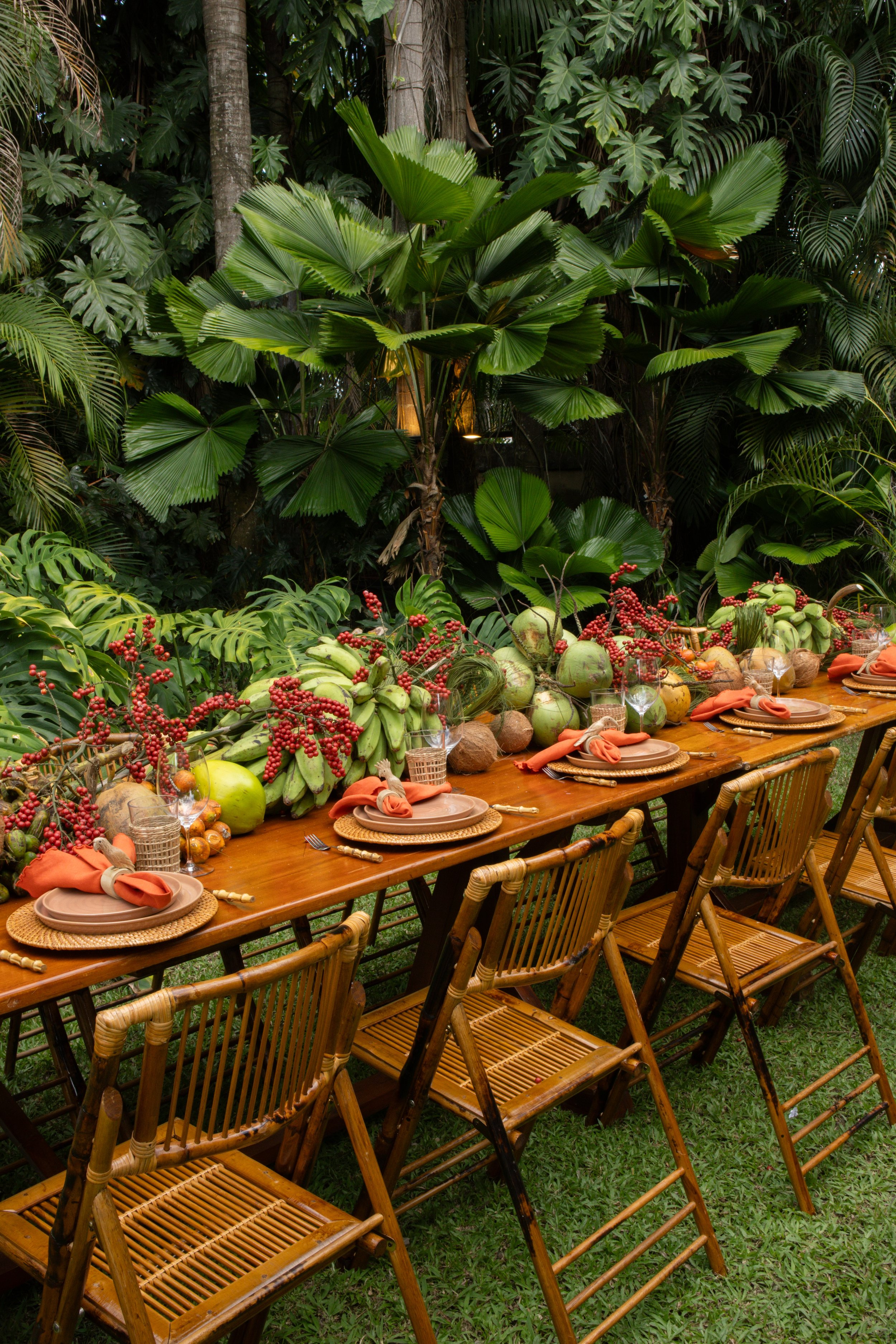 Mesa larga decorada con frutas tropicales, platos, vasos y utensilios, rodeada de una vegetación exuberante con muchas plantas verdes grandes.