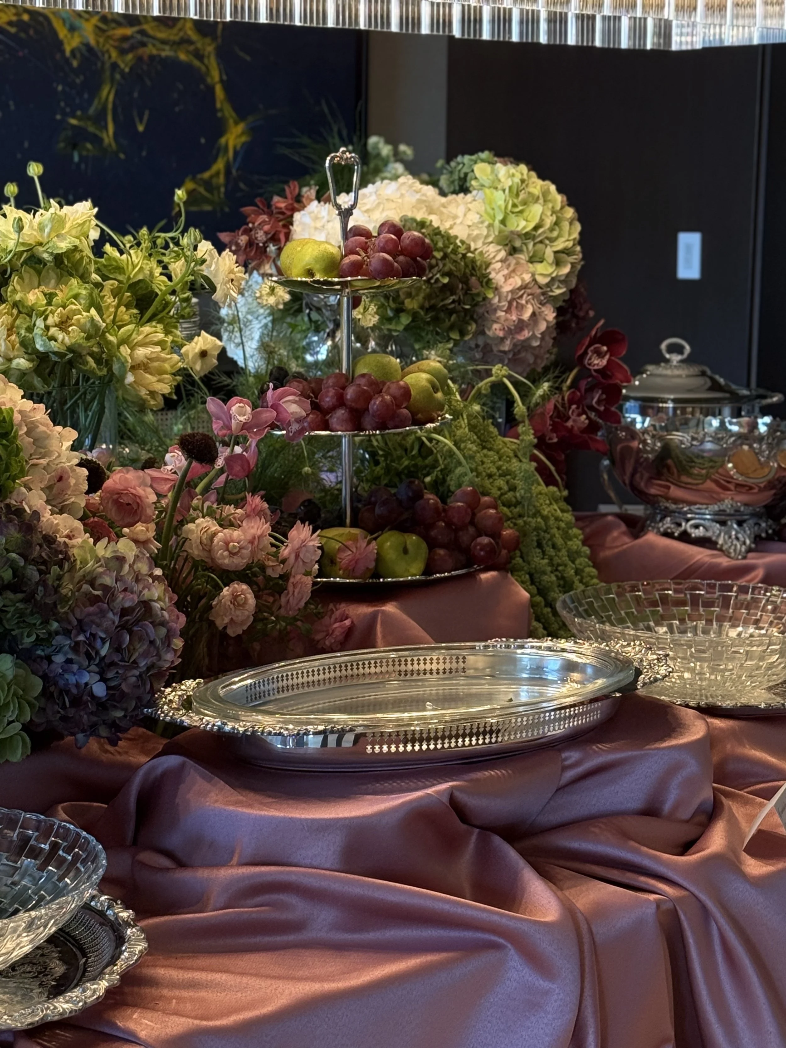 Arreglo floral con flores de diferentes colores, frutas en un plato de plata, y una mesa cubierta con tela de raso en tonos rosas y morados.