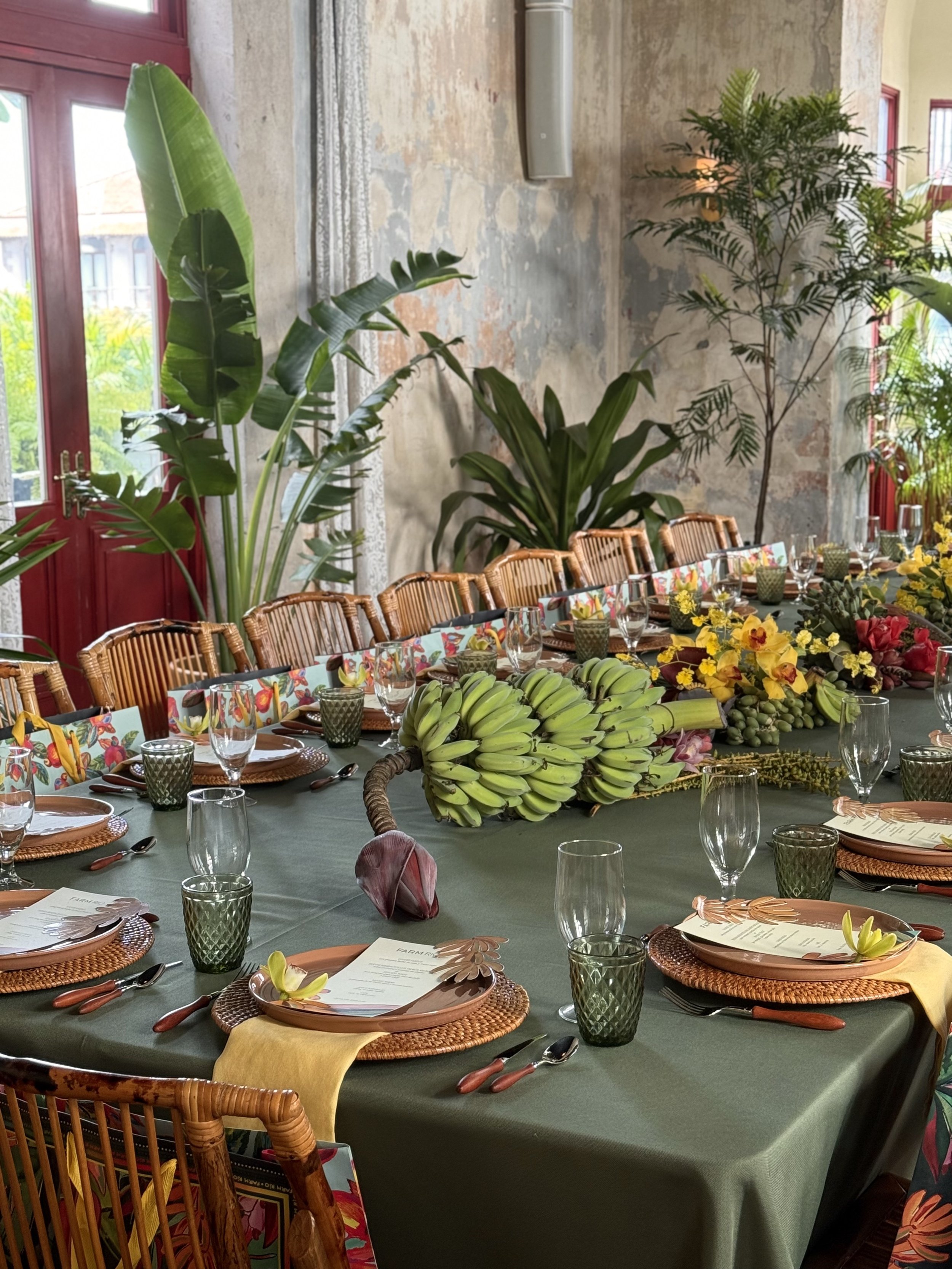 Mesa decorada con frutas, flores y vasos en un ambiente con plantas verdes y paredes de piedra, lista para una comida o evento.