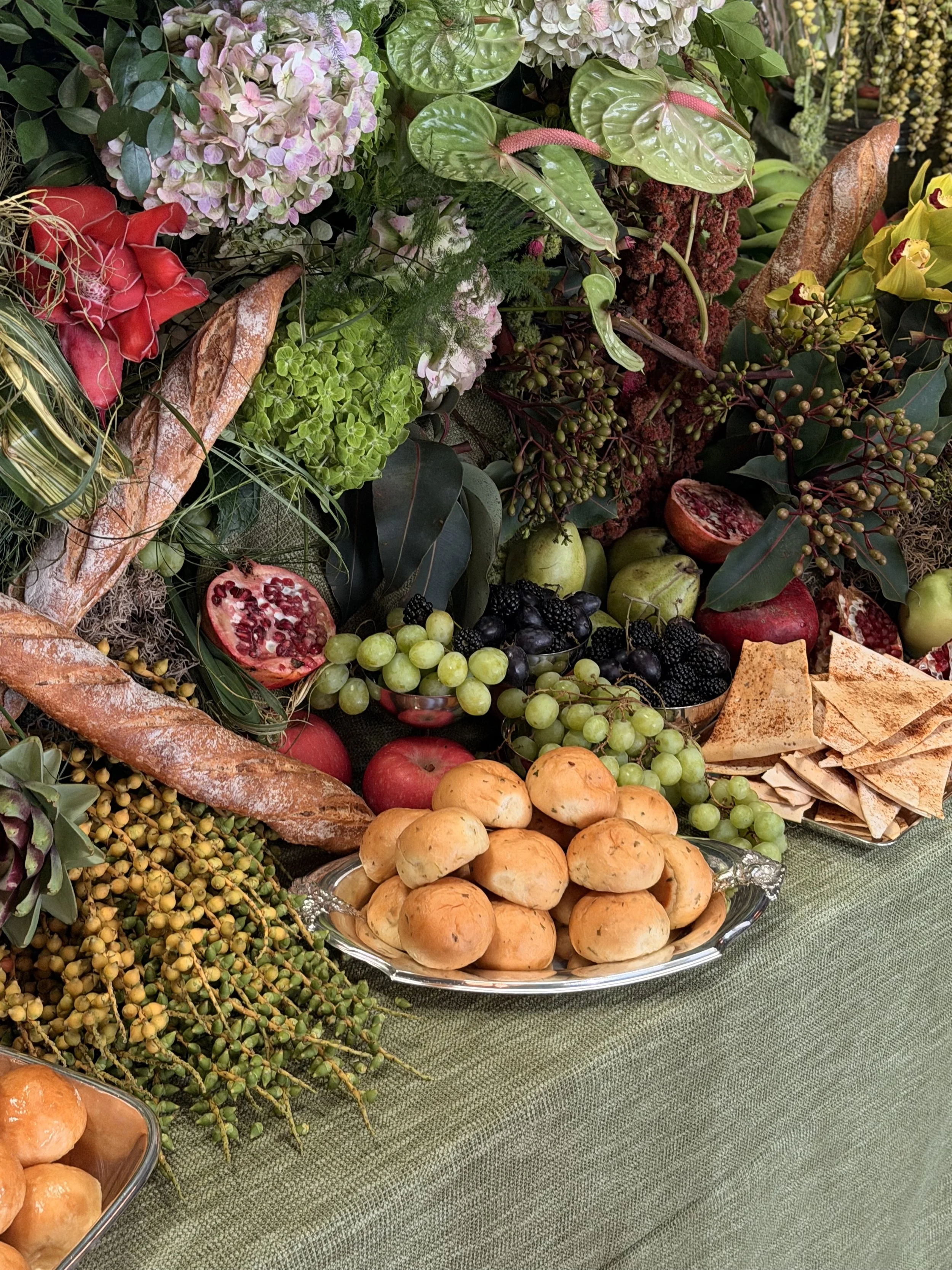 Una variedad de frutas y flores en un arreglo, incluyendo uvas verdes, uvas negras, granadas cortadas, manzanas, duraznos, panecillos, y diferentes tipos de flores y hojas verdes.