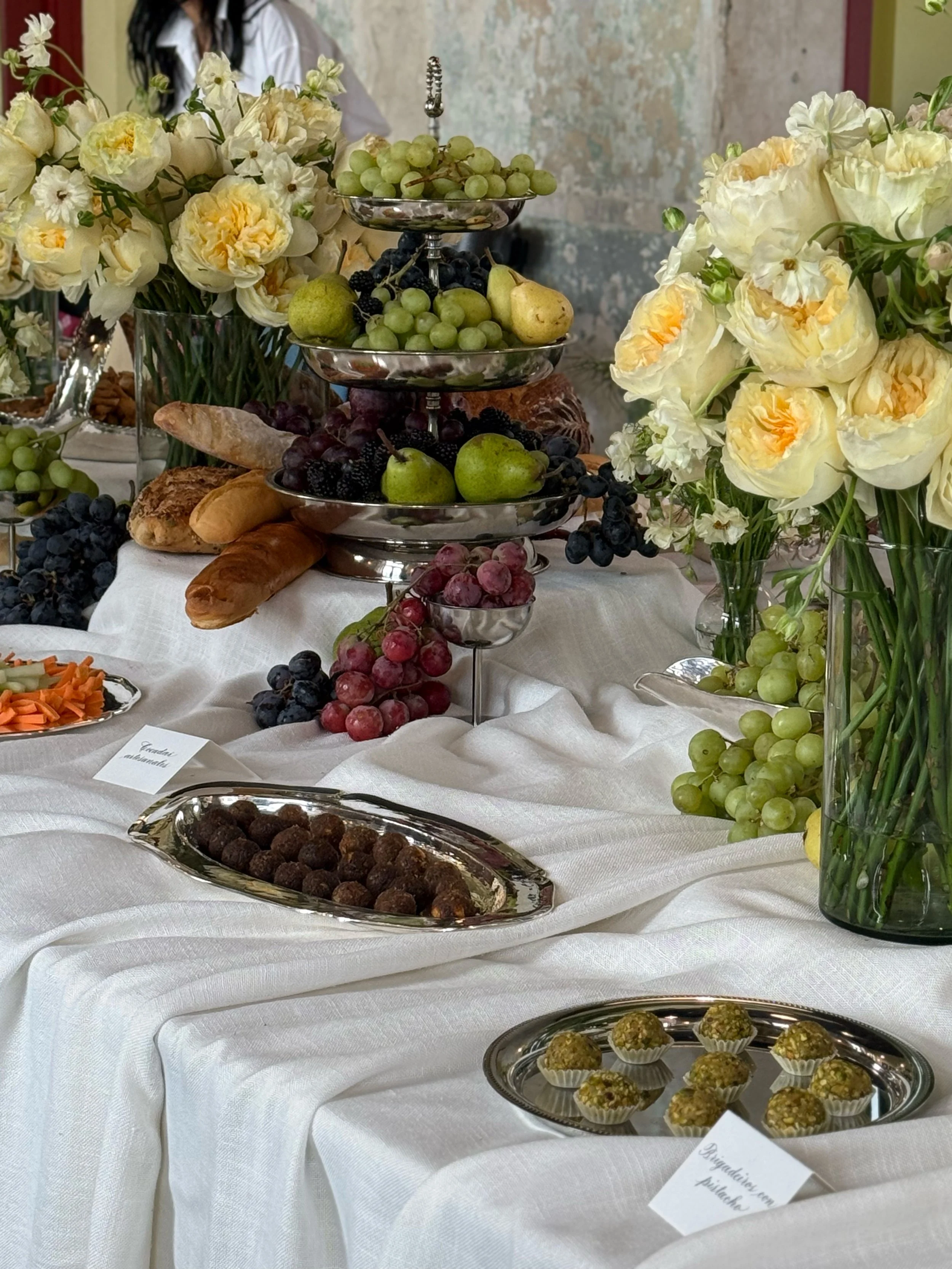 Mesa con flores blancas y amarillas, frutas como uvas verdes, moradas, peras y ciruelas, panes, y dulces en recipientes de plata y cristal, en un fondo con pared envejecida.