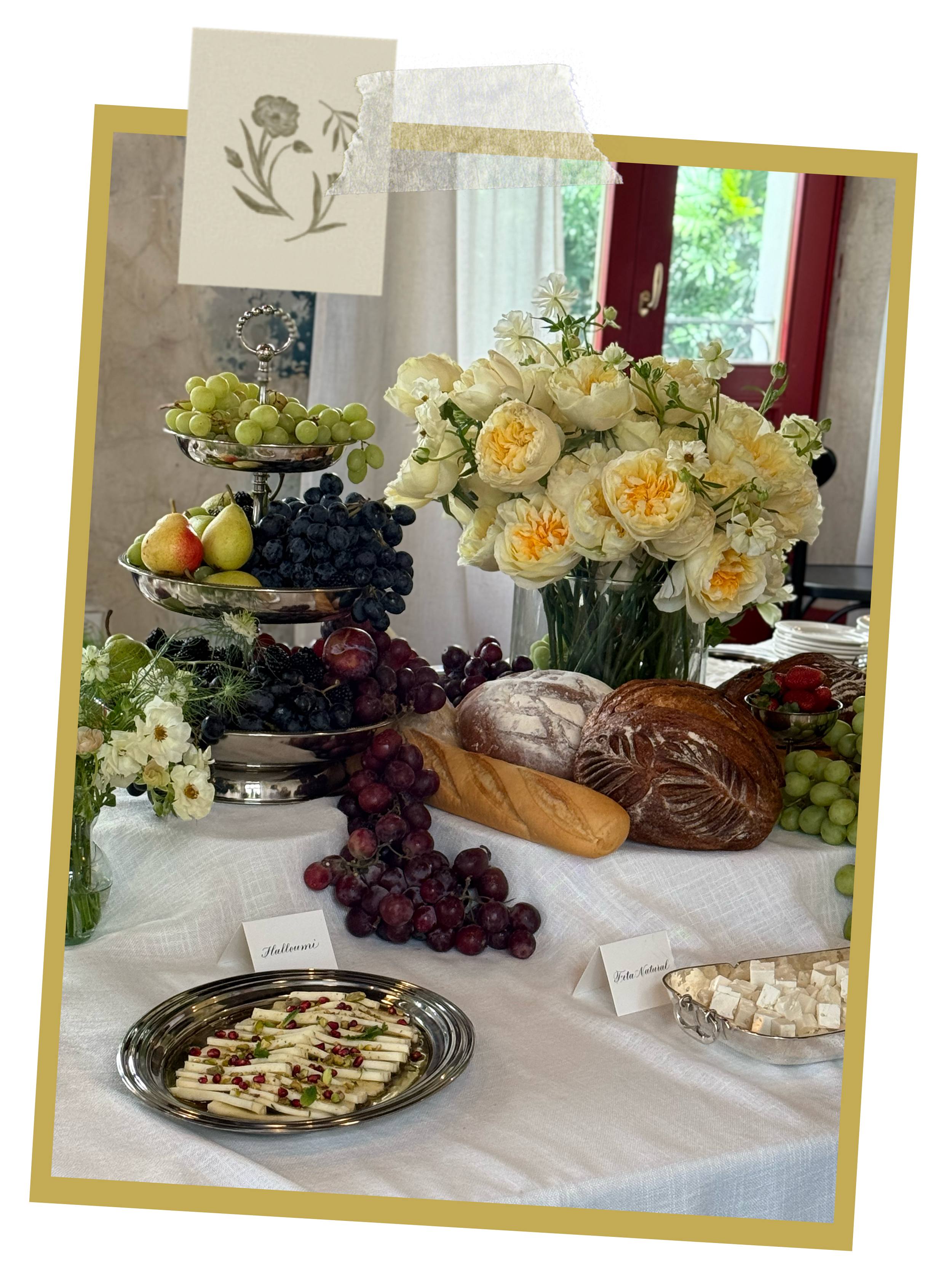 Mesa con variedad de frutas, pan, queso y flores decorativas en un ambiente luminoso y acogedor.