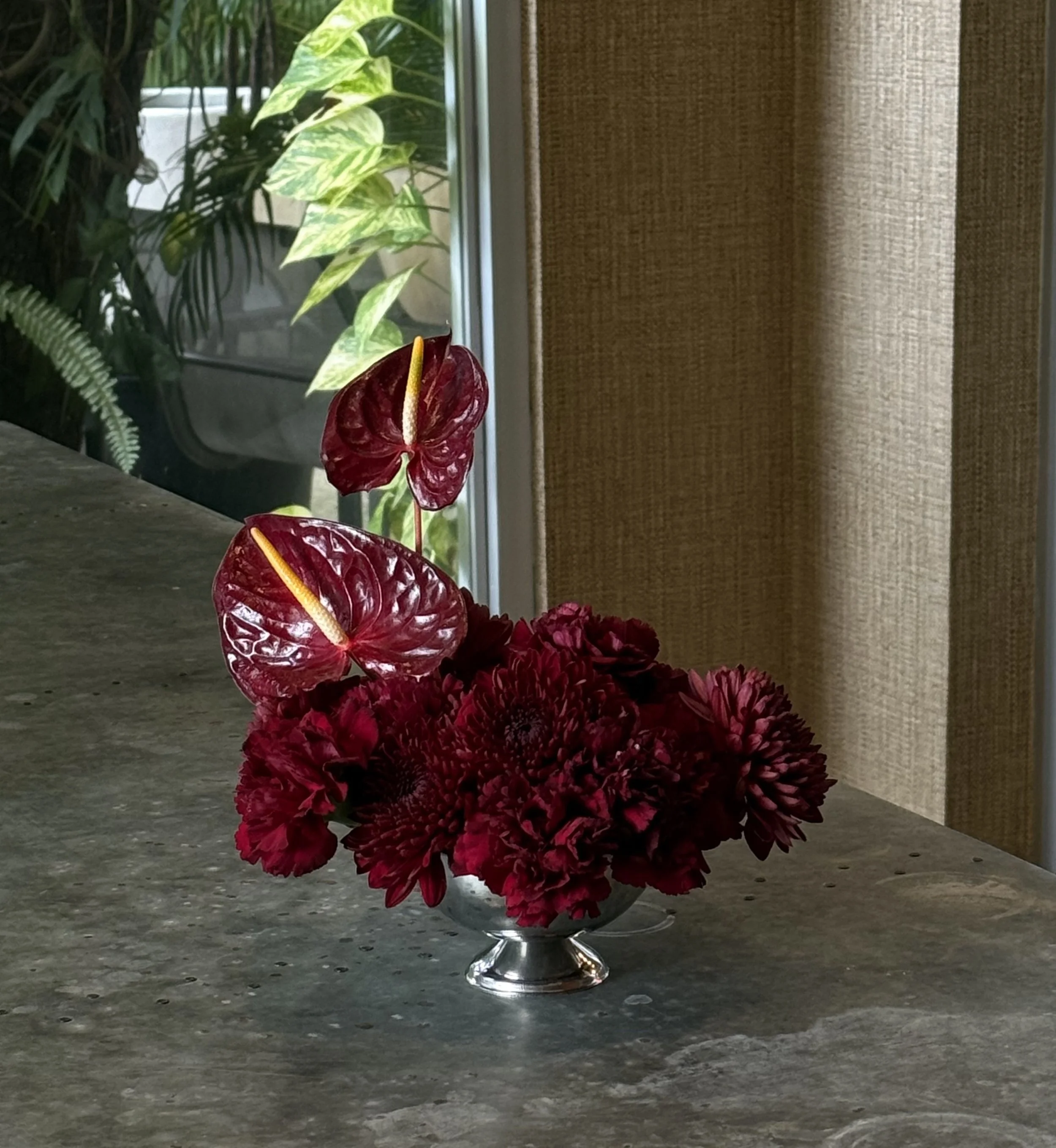 Florero con flores rojas y verdes, colocado sobre una mesa de piedra junto a una ventana con plantas verdes y cortinas beige.