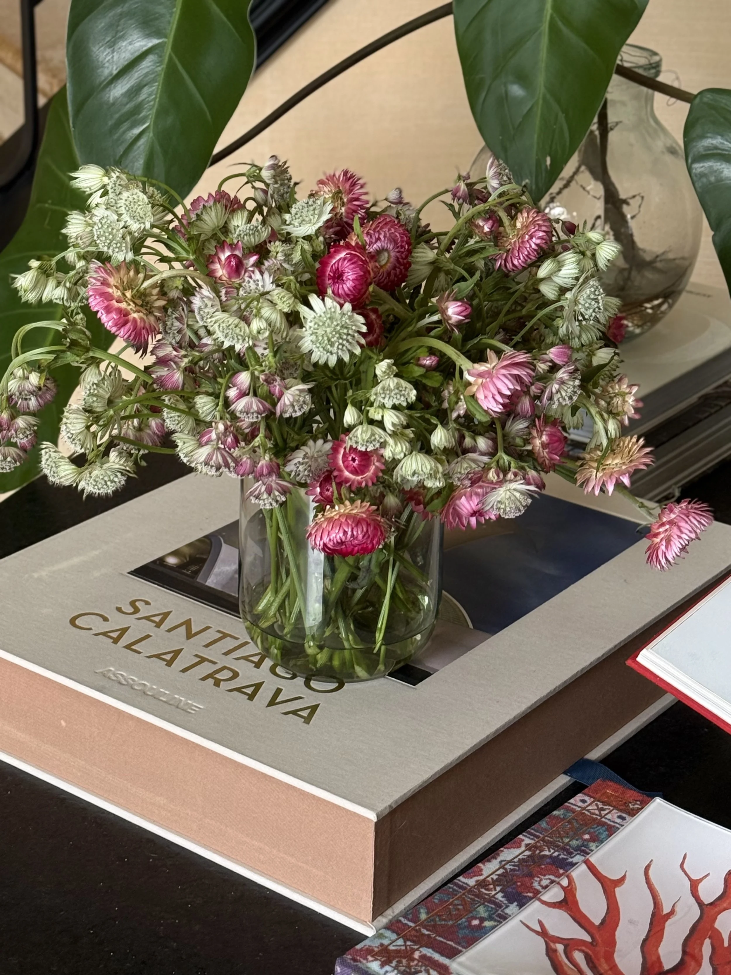 Ramo de flores variadas en un jarrón transparente, sobre una base con texto en español. Fondo con otras decoraciones y libros.