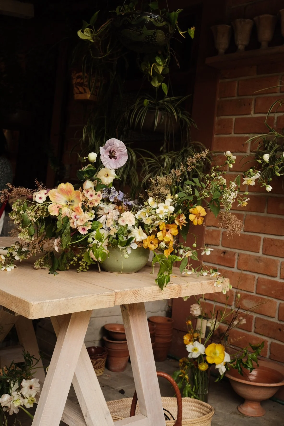 Arreglo floral con flores de colores en un recipiente en una mesa de madera en un espacio con paredes de ladrillo y plantas colgantes.