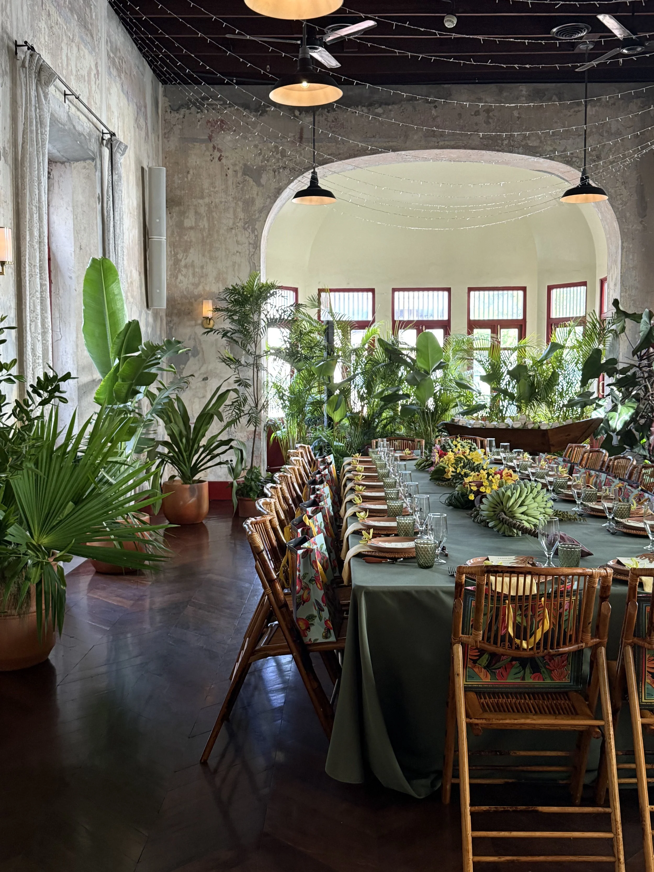 Mesa larga con decoración floral y vajilla en un restaurante con plantas grandes y paredes de estilo rústico, iluminación suave y ventanas grandes con marcos de madera.