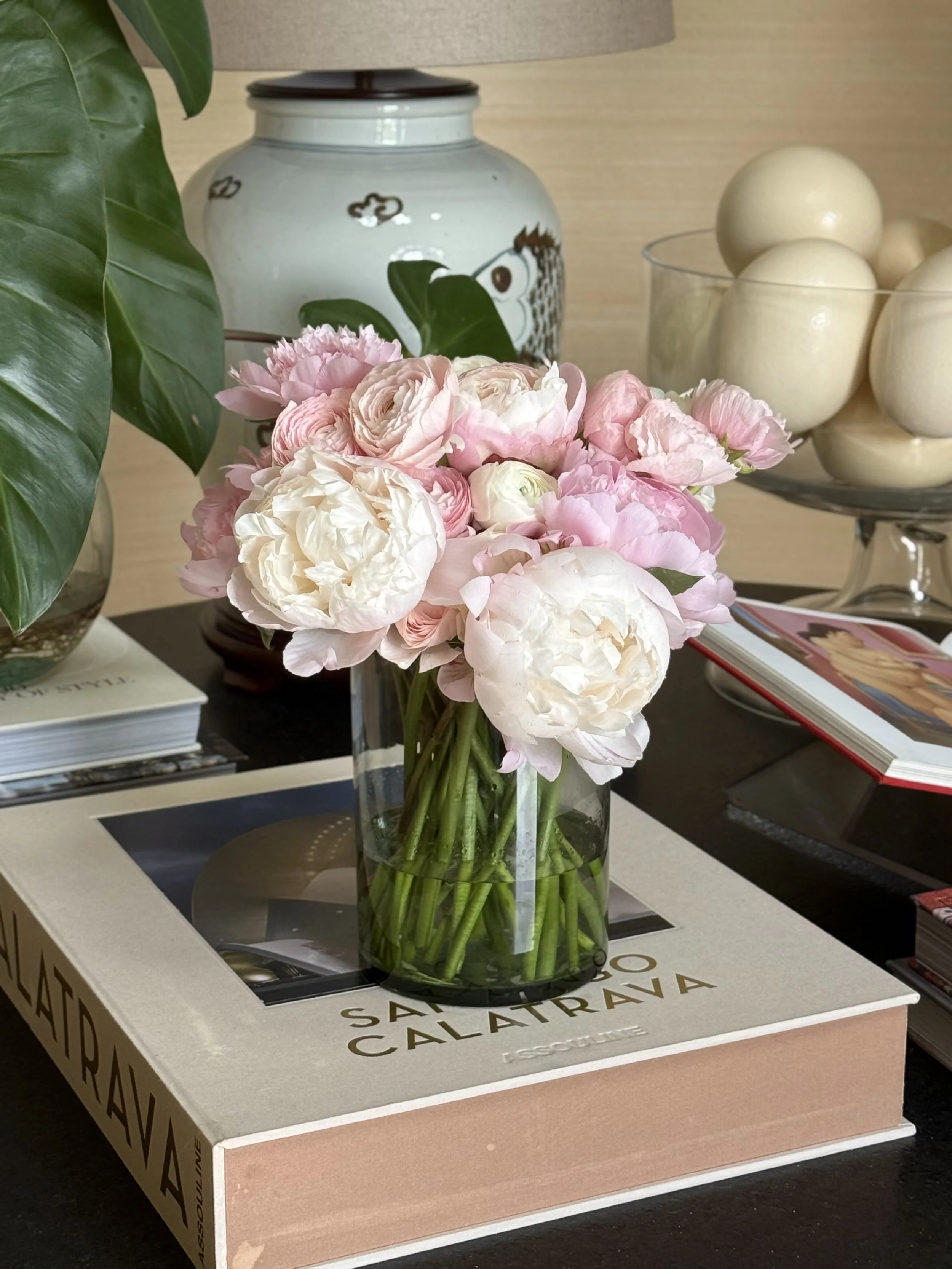 Ramo de flores rosas y blancas en un florero de vidrio, jarrón decorativo con dibujo de un pez, caja con huevos blancos, libro y revista en una mesa de madera