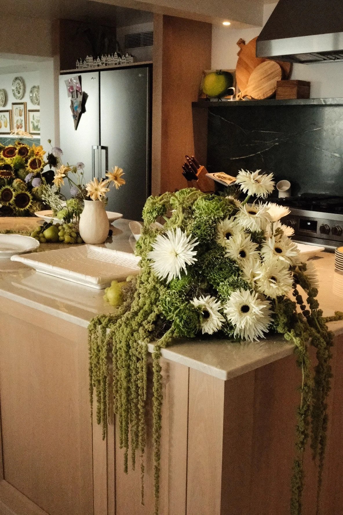 Mesa de cocina decorada con arreglos florales blancos y verdes, con platos blancos y una bandeja con frutas verdes, en un ambiente cálido con utensilios de cocina y estantes de madera en el fondo.