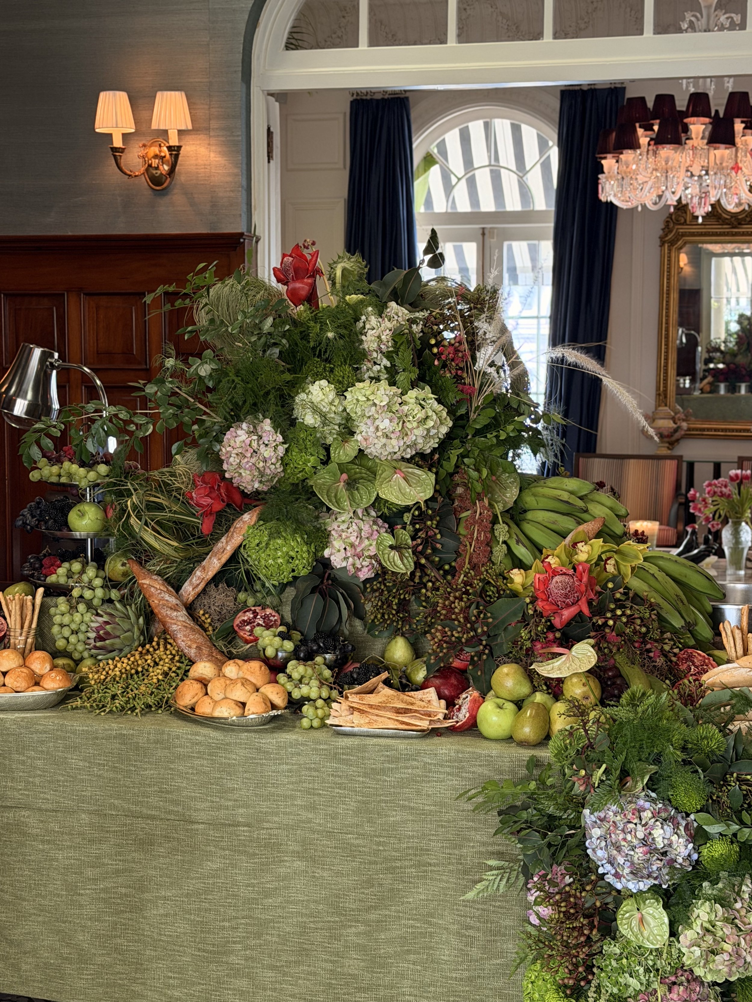 Una mesa decorada con una abundante variedad de flores y frutas, con un fondo interior de una habitación elegante con cortinas azules, un espejo dorado y un candelabro con luces.