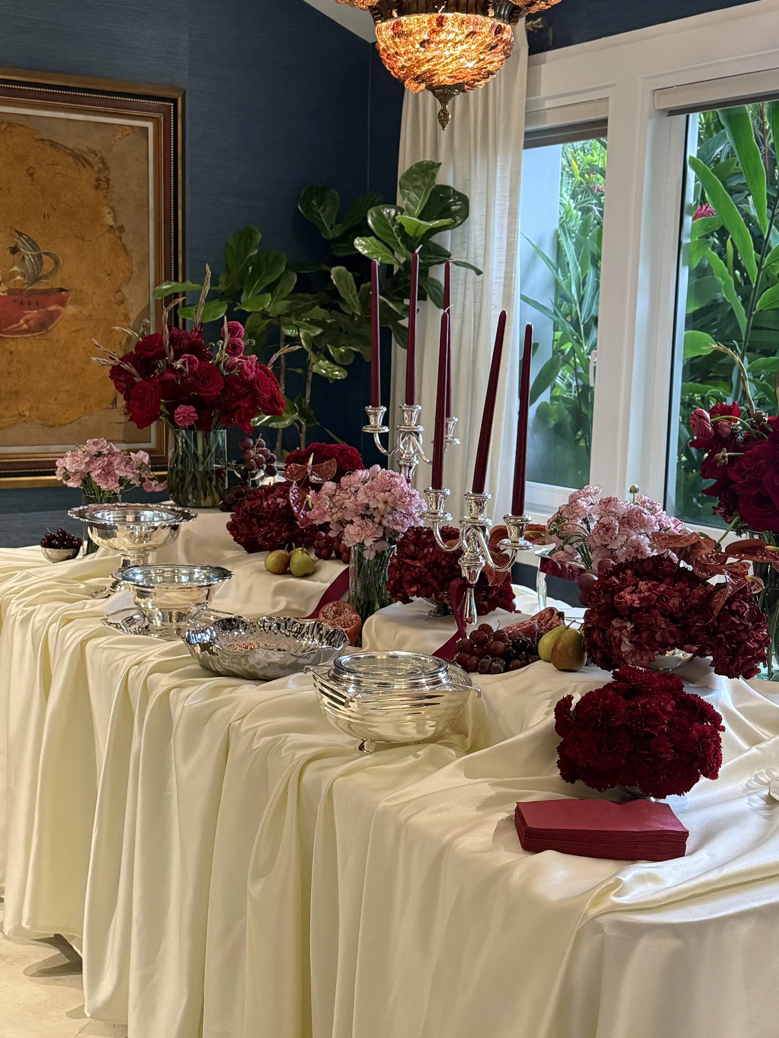 Mesa decorada con flores rojas y rosadas, candelabros con velas moradas, frutas y vasos de plata en un entorno interior con cortinas blancas y ventanas que muestran plantas verdes.
