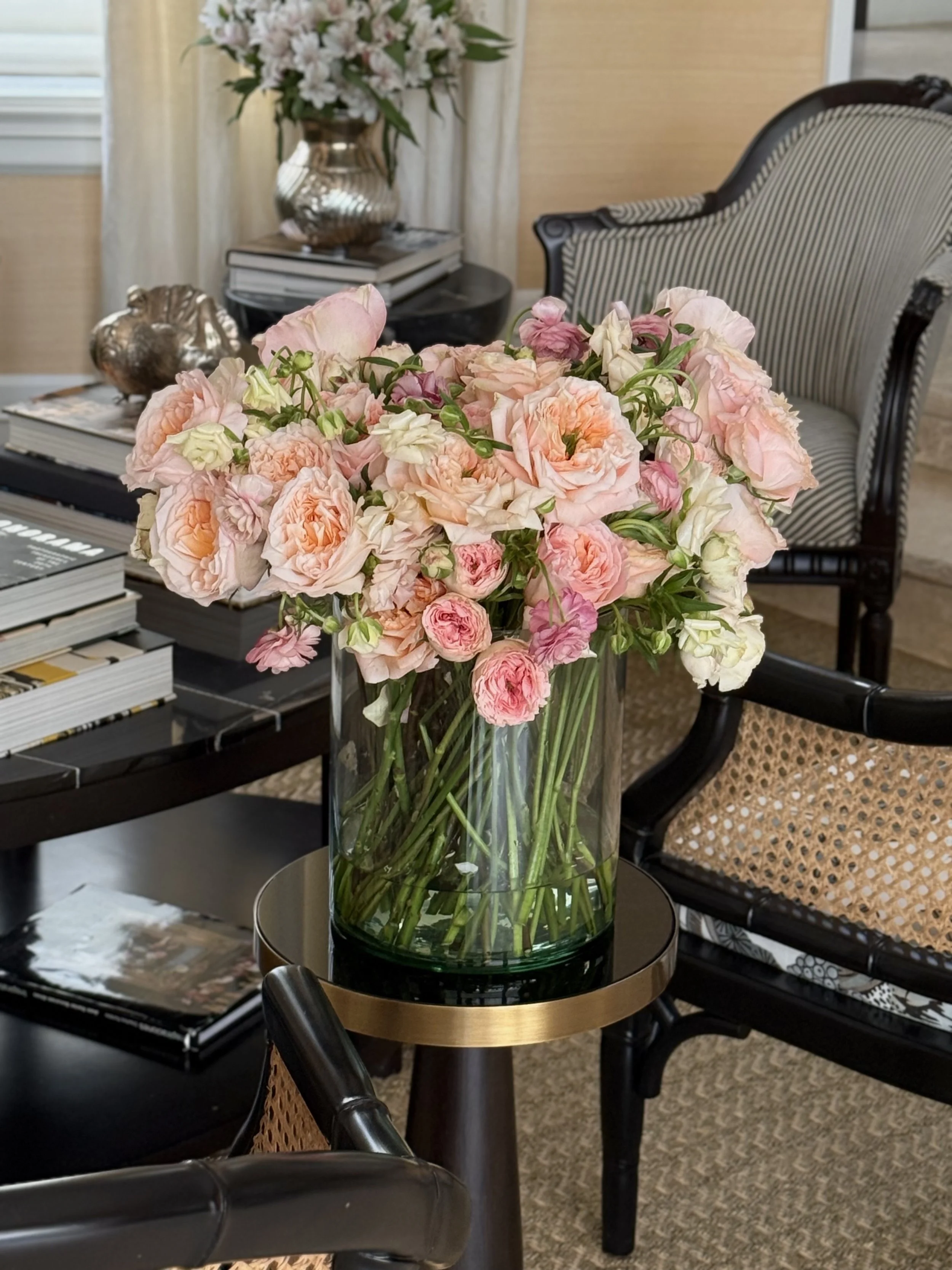 Ramo de flores rosas en un florero de vidrio en una mesa redonda de madera. En el fondo, una silla con respaldo de madera y ratán y un rincón decorado con libros y objetos plateados, y una planta con flores blancas en un jarrón.