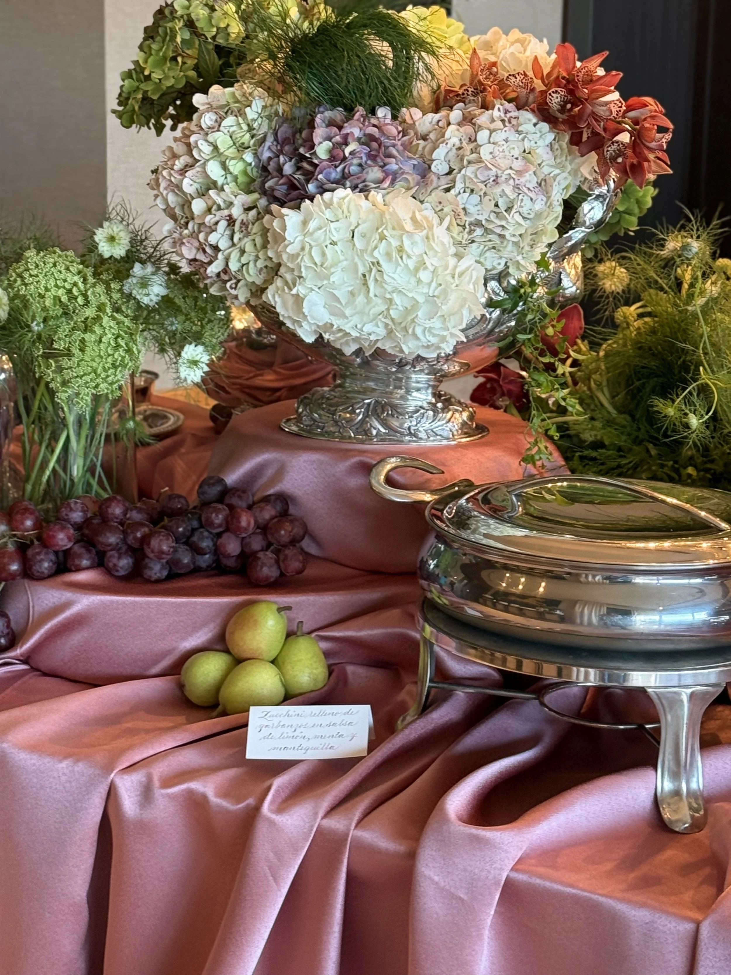 Un arreglo de flores en un bol de plata con uvas, manzanas verdes y una etiqueta con texto en español.