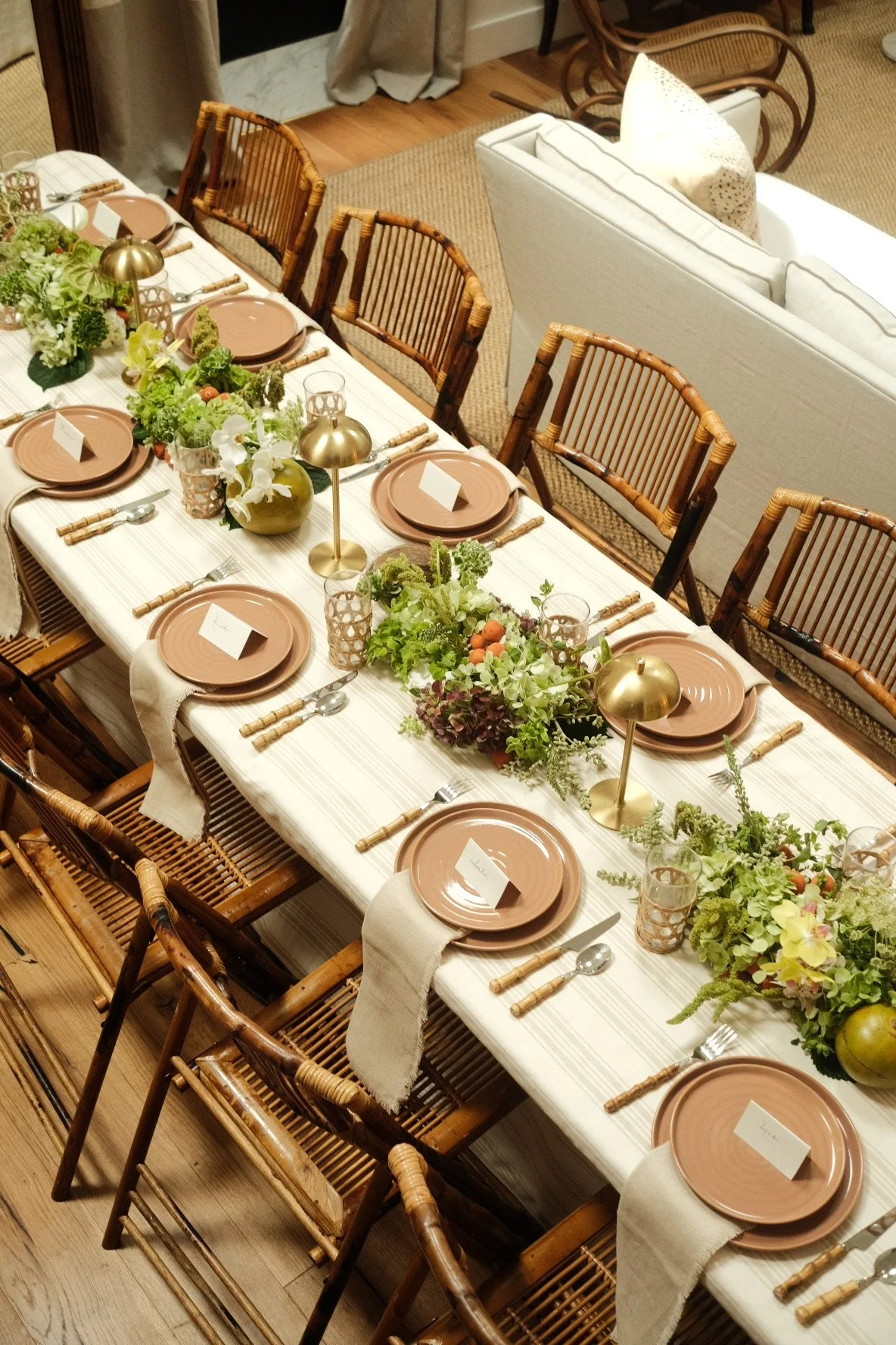 Mesa decorada para comida con platos rosas, cubiertos de madera, centros de flores verdes y dorados, y manteles blancos con rayas suaves.