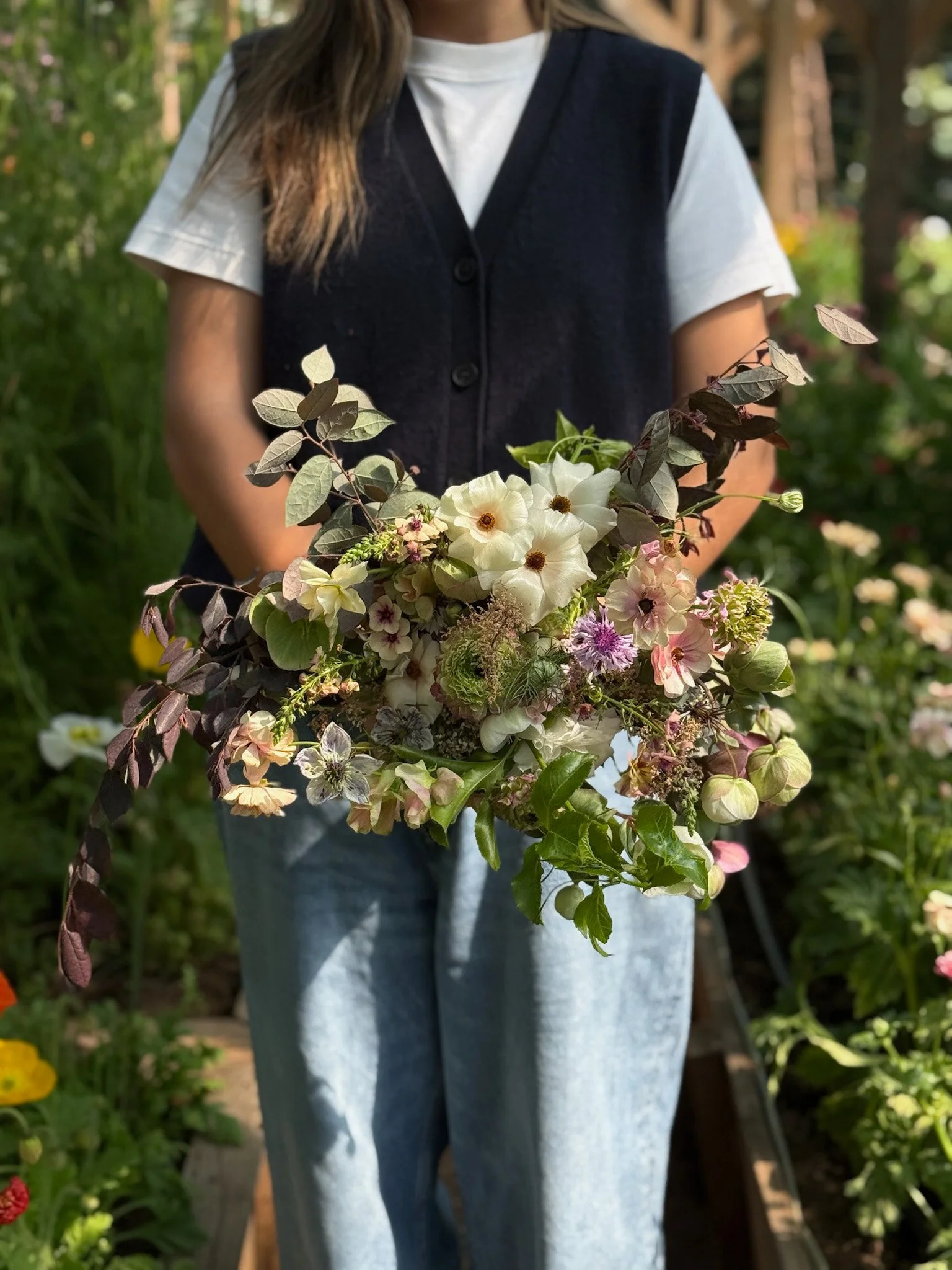 Persona sosteniendo un ramo de flores en un jardín en exteriores, con árboles y plantas de fondo.