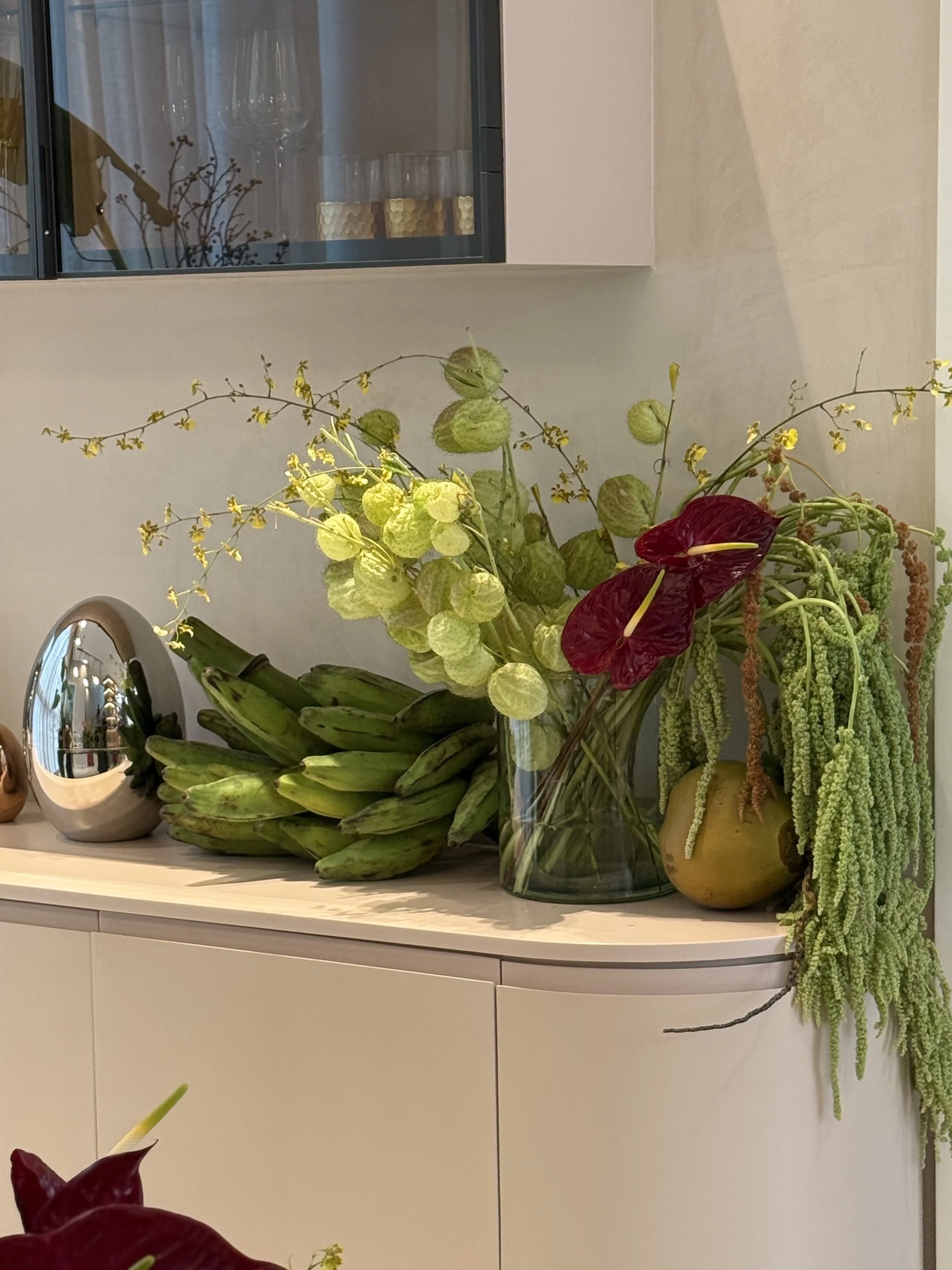 Arreglo de flores en una mesa blanca, incluyendo plantas verdes y flores de colores rojos y amarillos, con vasos y objetos decorativos al fondo.