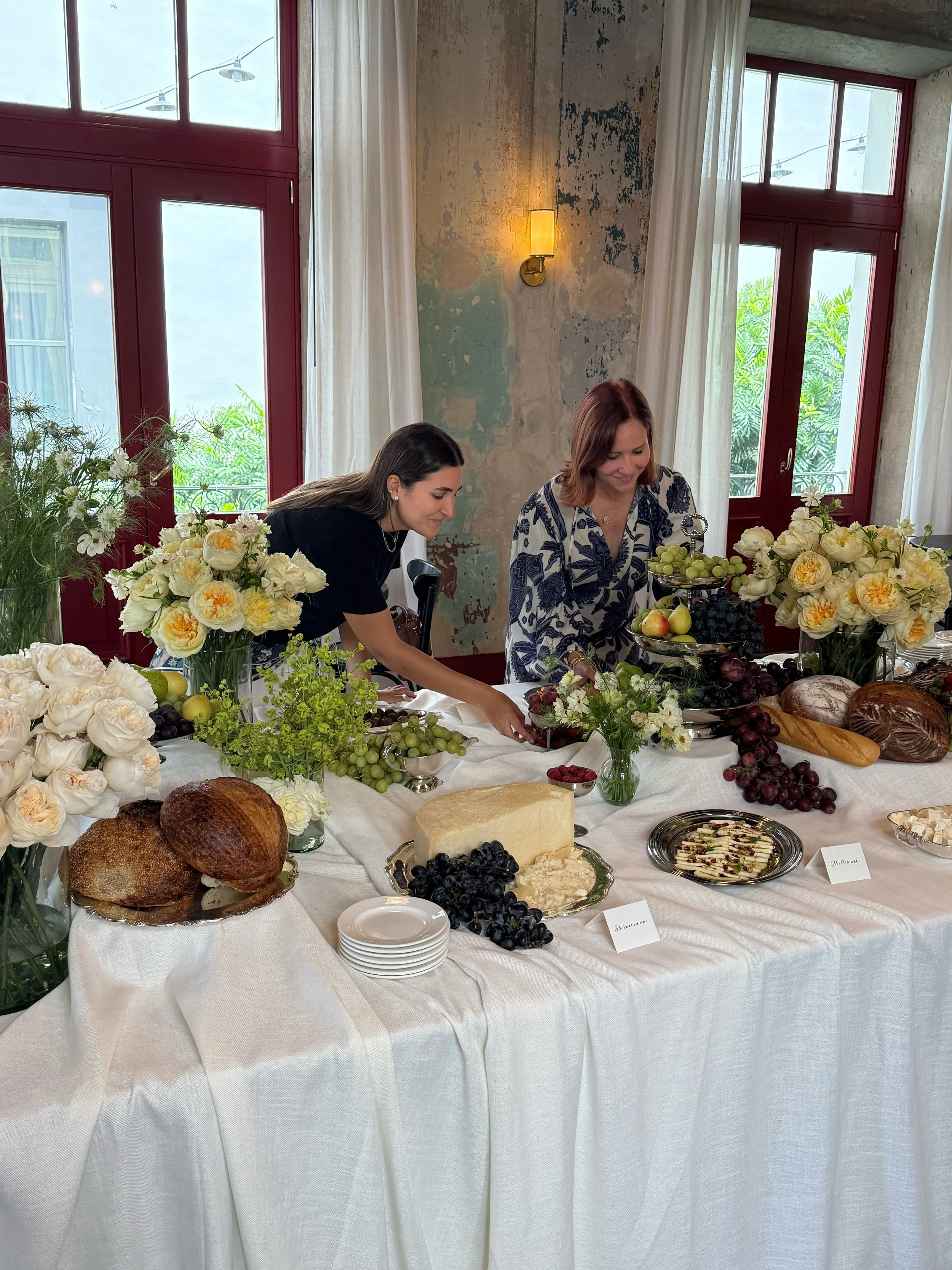 Mesa decorada con flores, panes, quesos, frutas y otros productos, en un ambiente con ventanas grandes y paredes envejecidas.