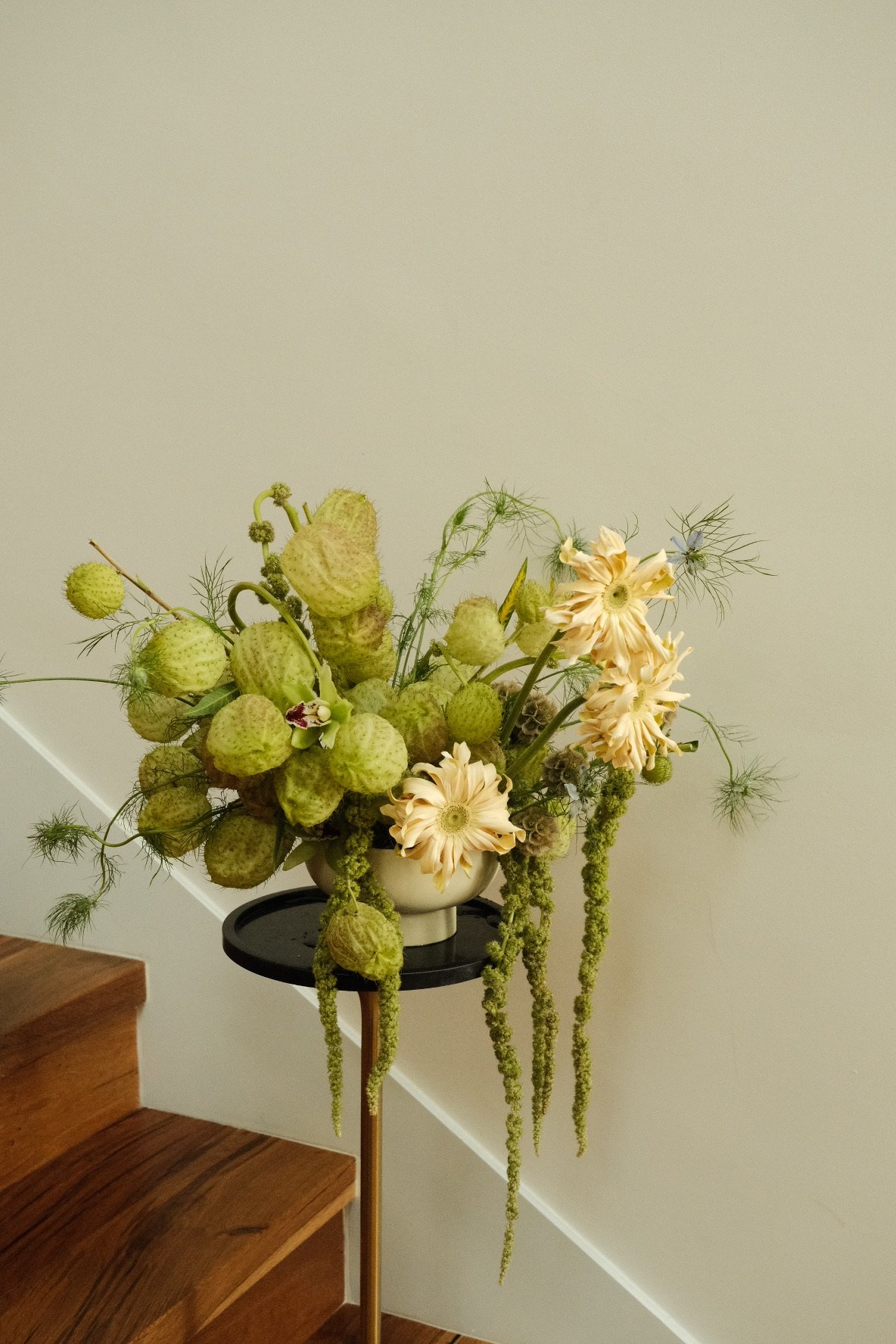 Ramo de flores secas y plantas en un florero en una mesa alta junto a escaleras de madera en un espacio interior.