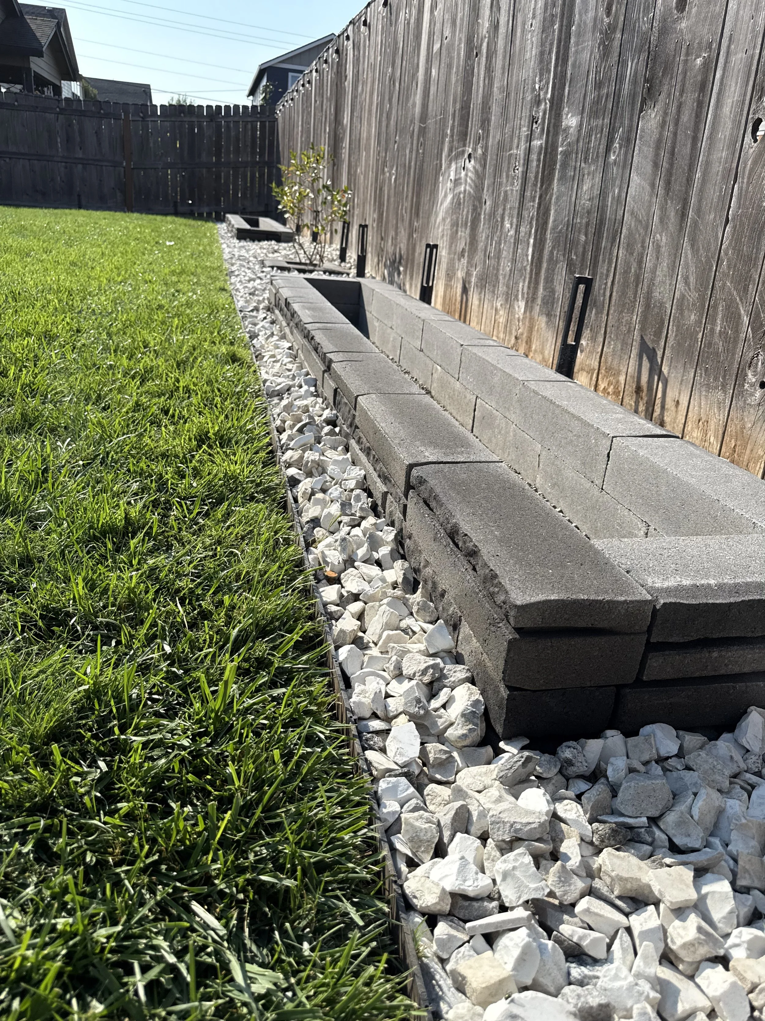 A backyard with a wooden fence, a small tree, a row of cinder blocks, and a line of white gravel alongside a strip of green grass.