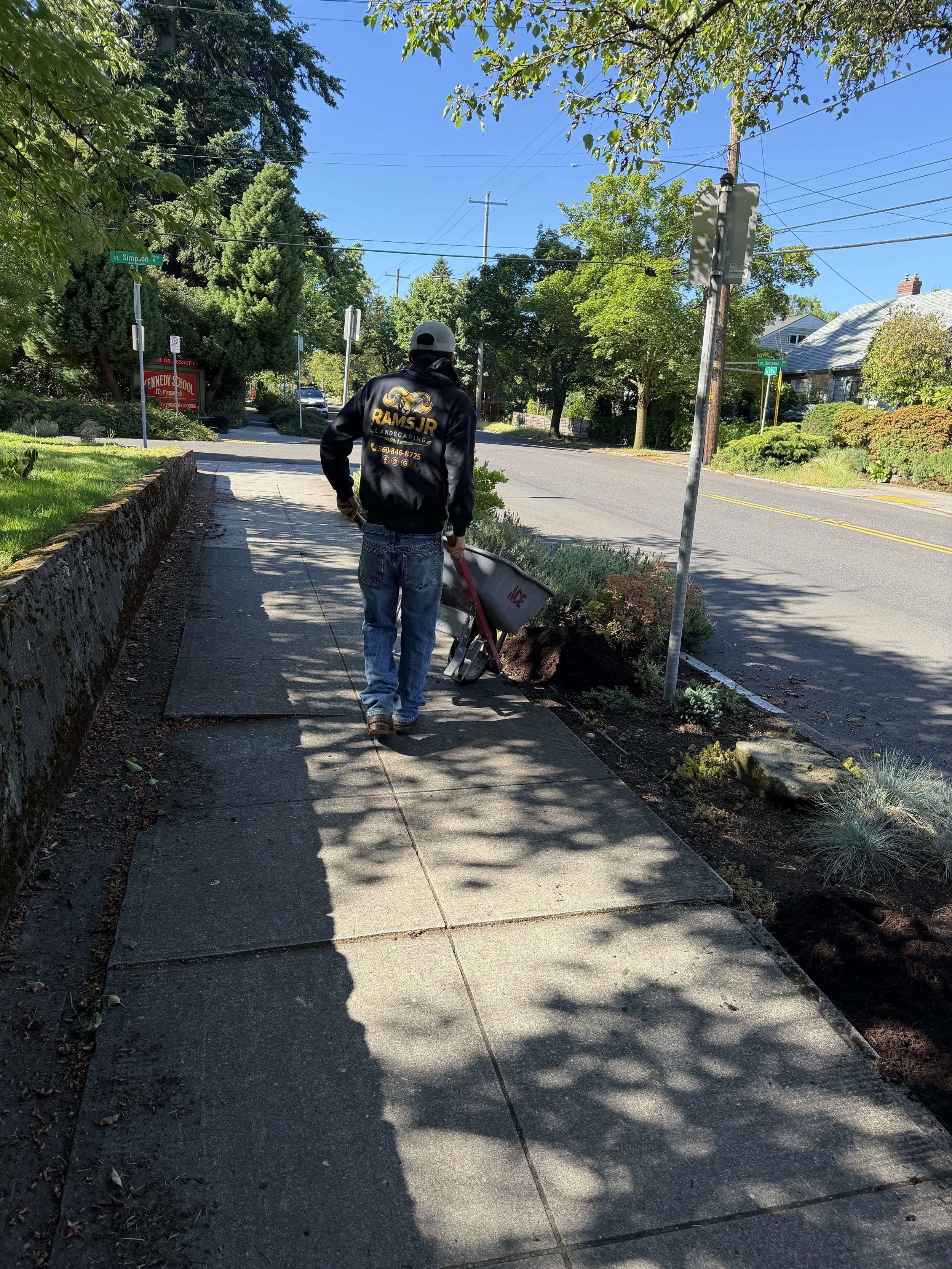 A person pushing a wheelbarrow along a sidewalk on a sunny day, with a row of trees, bushes, and houses in the background.