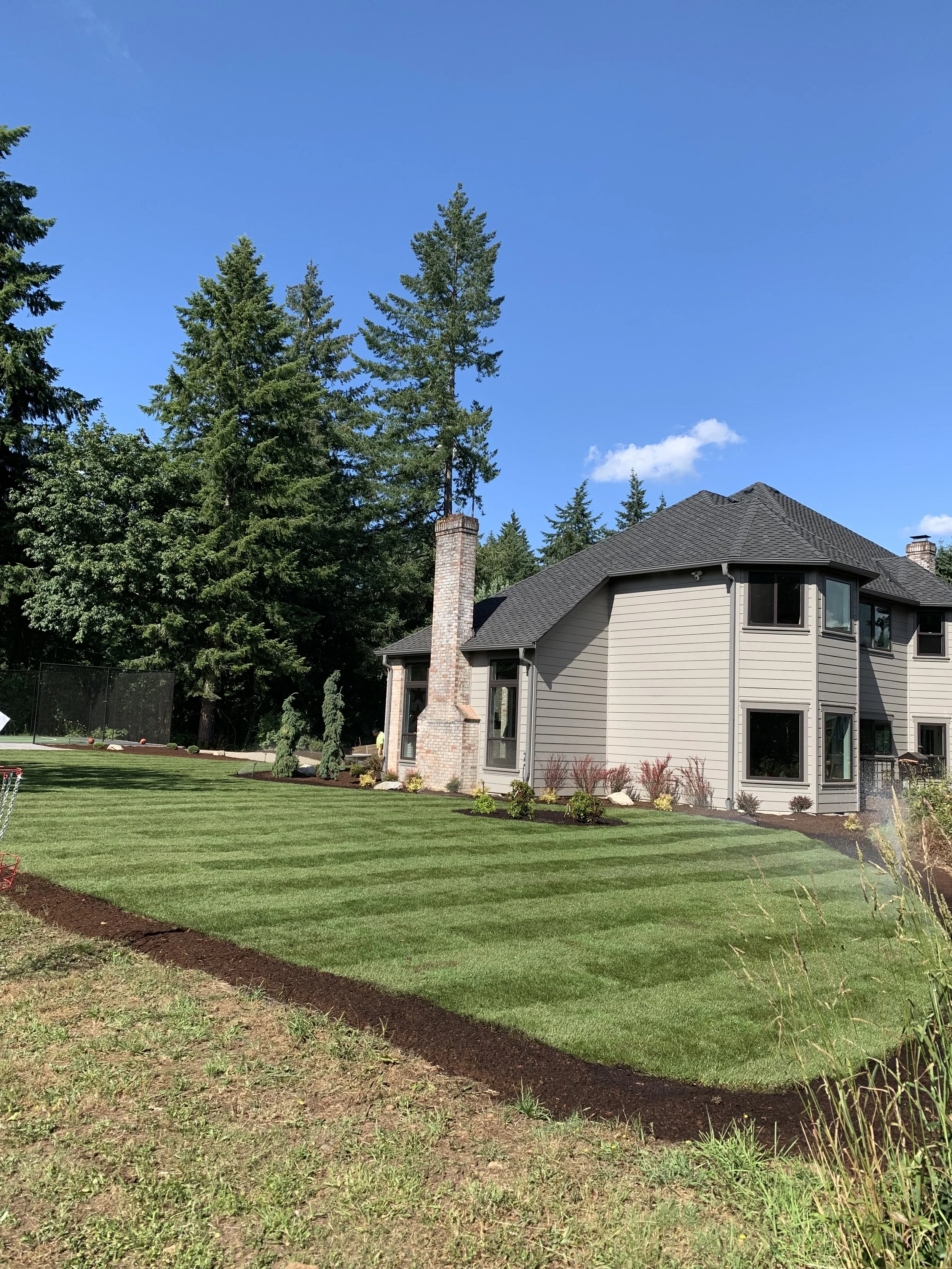 A backyard with a well-maintained green lawn, newly planted garden beds, and trees, with a house featuring a chimney and several windows, under a clear blue sky with a few clouds.