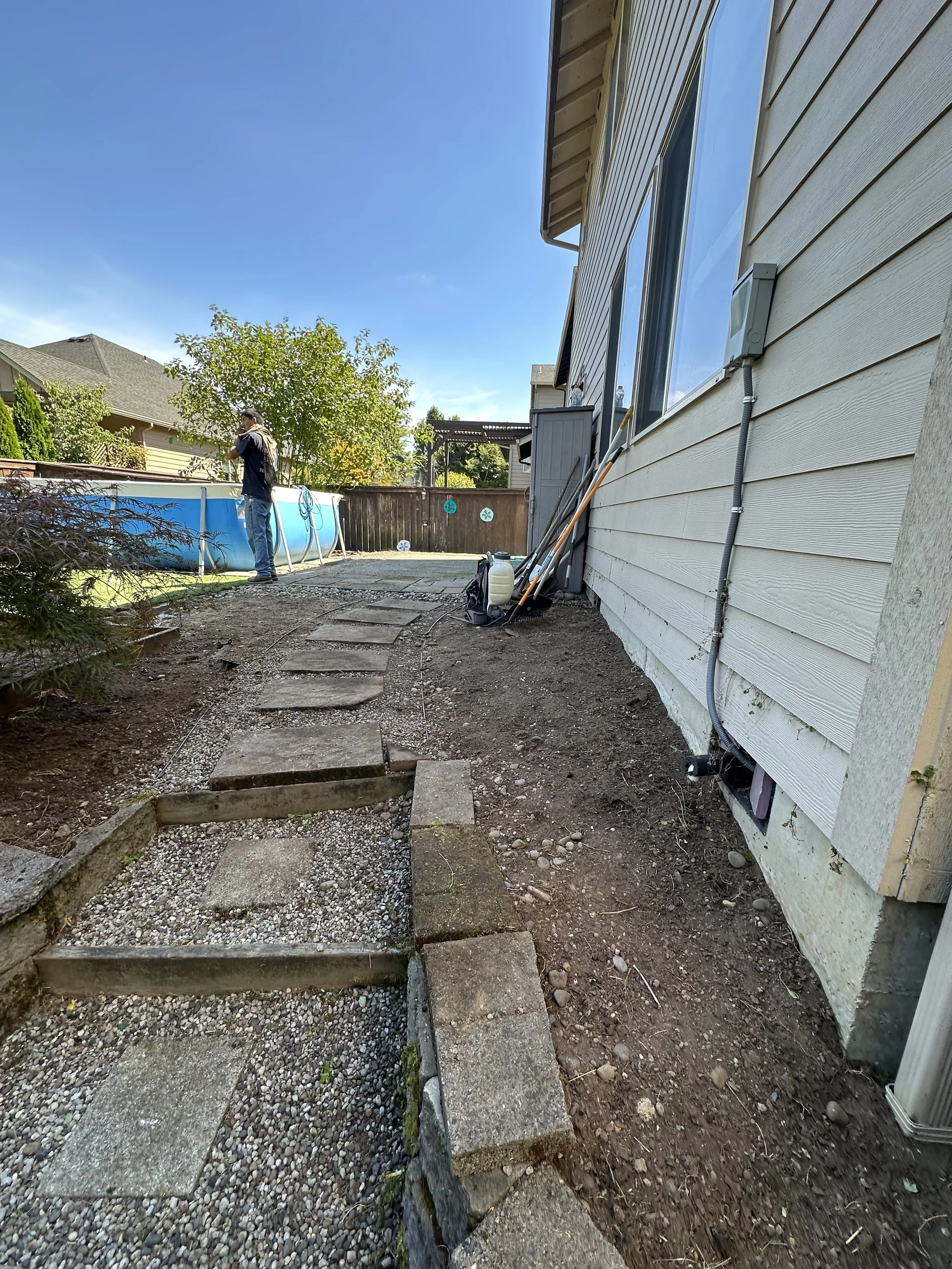 Backyard with a small above-ground pool, stepping stones, and a dirt pathway along the house wall. A person is standing near the pool, and there are trees and houses in the background.