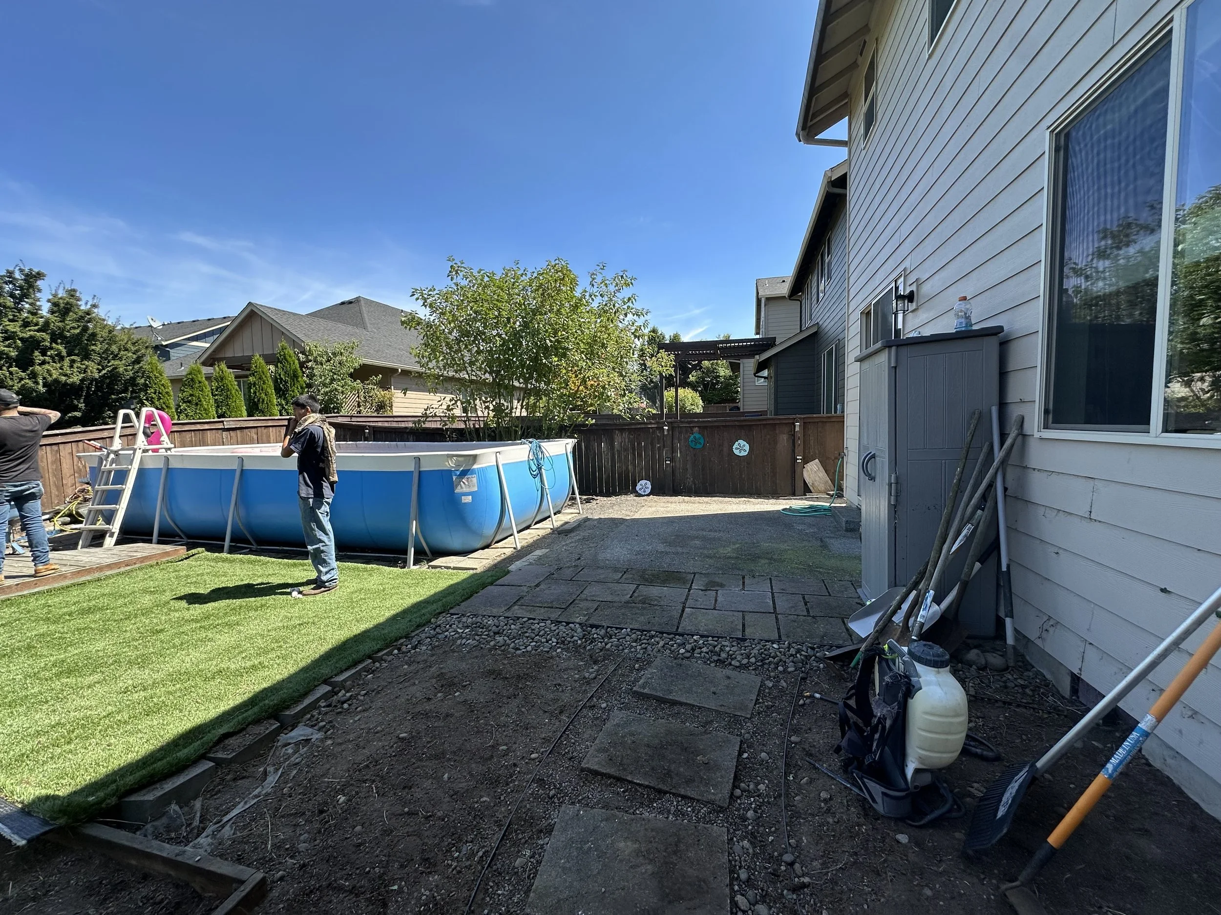 Backyard with a above ground swimming pool, people working on yard, partially finished landscaping, house with beige siding, and clear blue sky