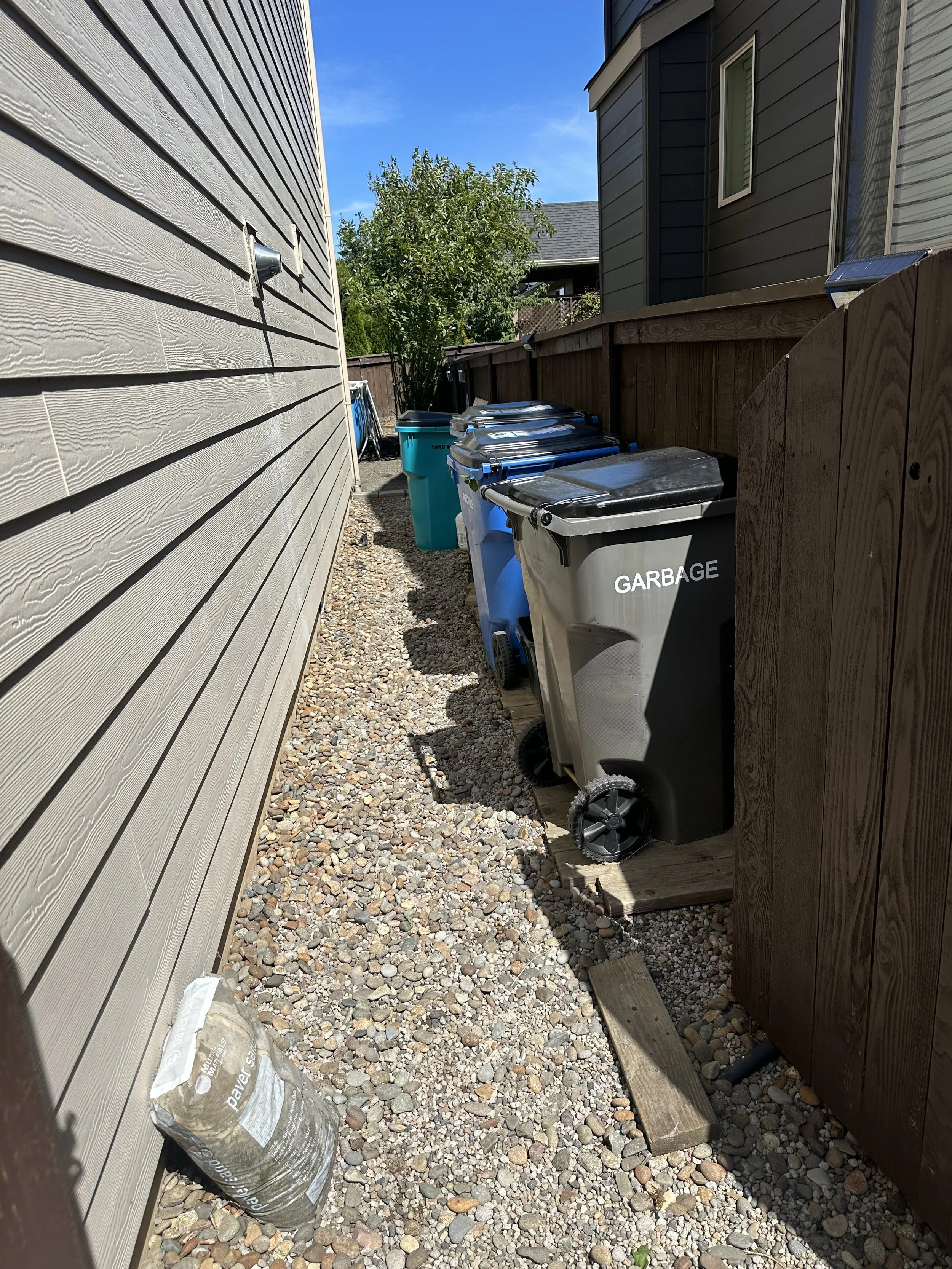 Backyard alley with four trash bins lined up along a wooden fence, a bag of gravel on the gravel ground, and a house on each side.