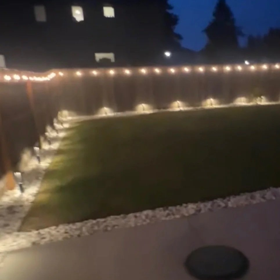 A backyard at night with string lights along a wooden fence and spotlights illuminating a grass yard.