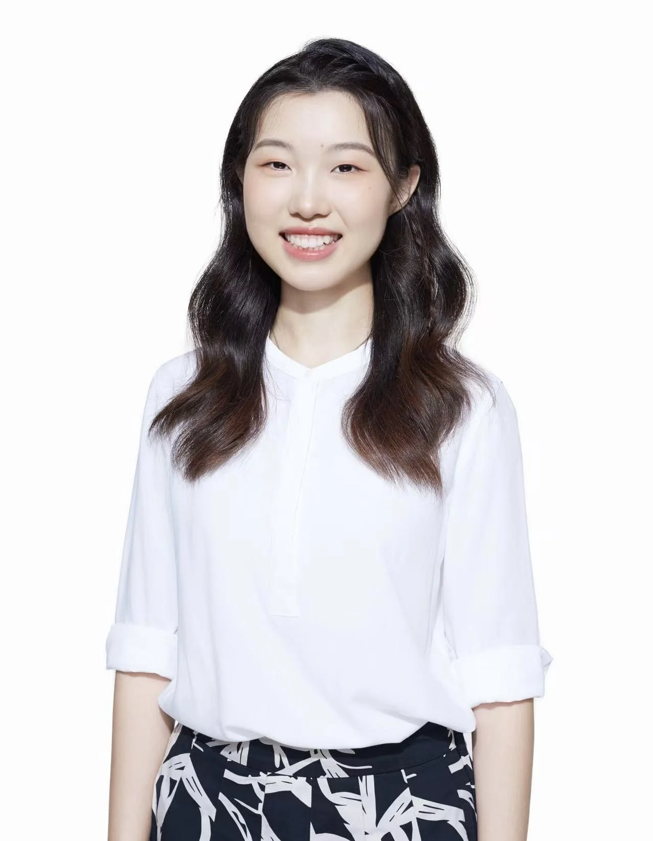 A young woman with long, wavy black hair smiling, wearing a white blouse and patterned black and white pants, against a plain white background.