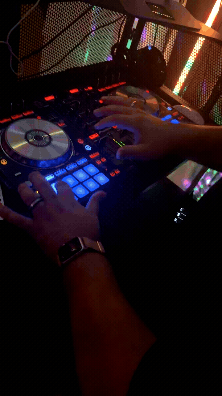 A person using a DJ controller with colorful buttons and a turntable, in a dimly lit setting with multicolored LED lights.