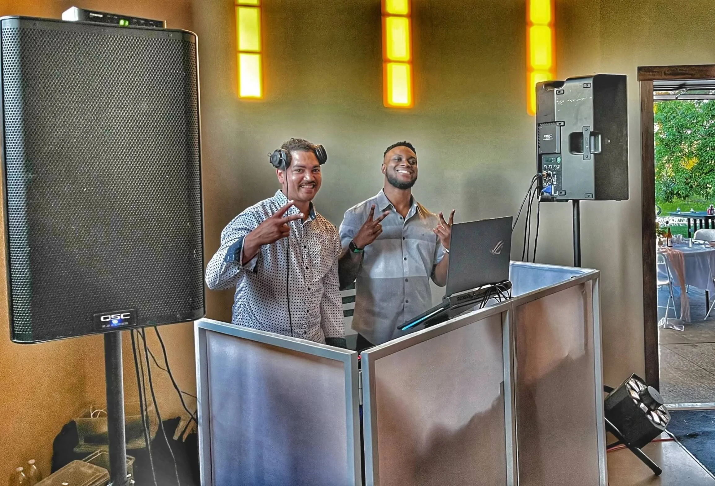 Two smiling men standing behind DJ equipment, posing with peace signs, in a room with large speakers and yellow lighting panels.