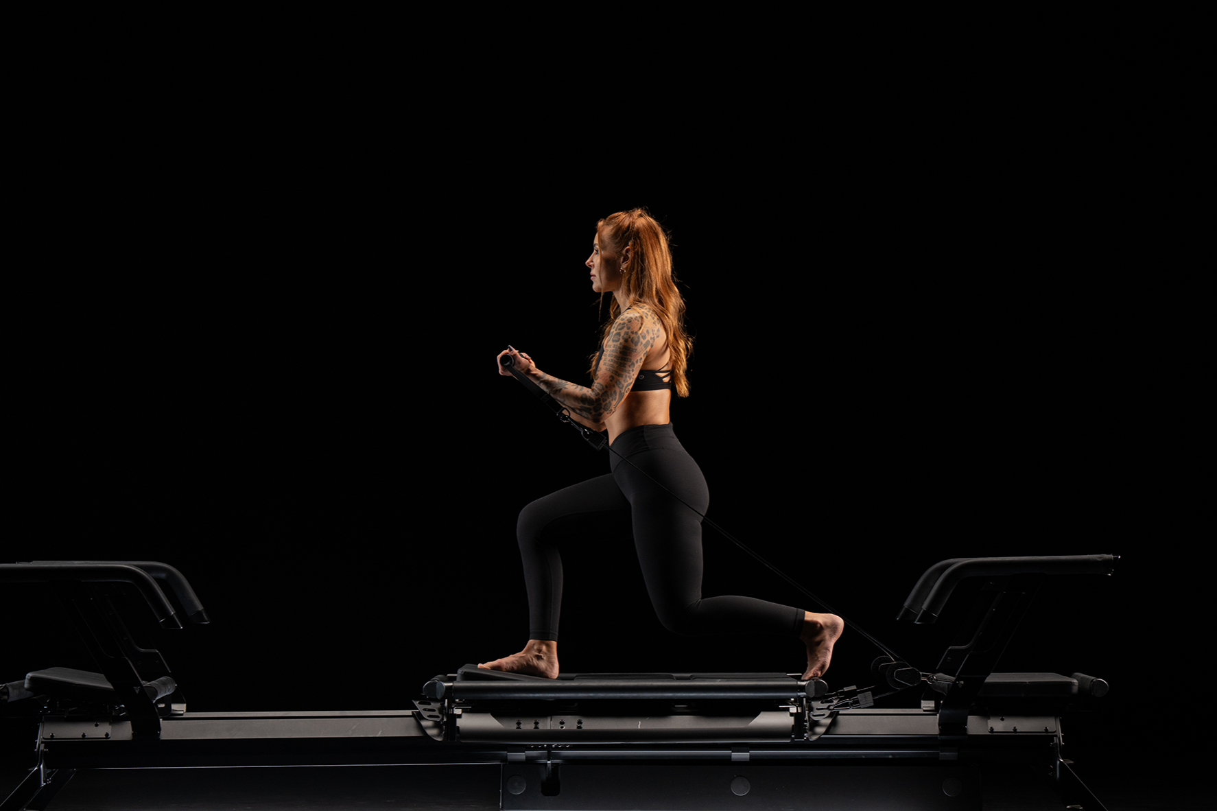 A woman with red hair and tattoos wearing black workout clothes kneels on a Pilates reformer machine in a dark studio, illuminated by a single light underneath.