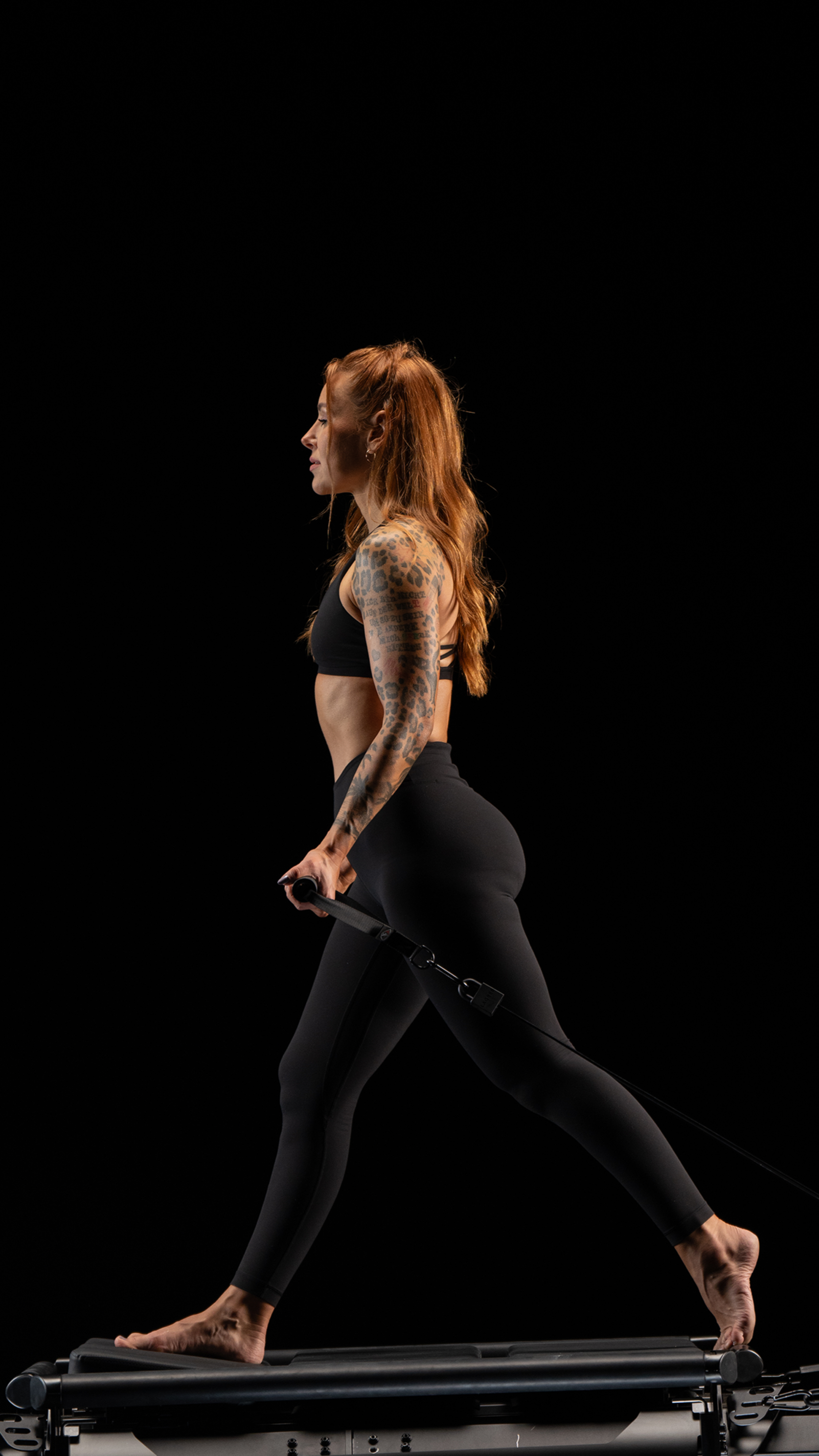 A woman with tattoos on her right arm performs a move on a Lagree EVO RAMP in a dark studio, wearing black athletic clothing.